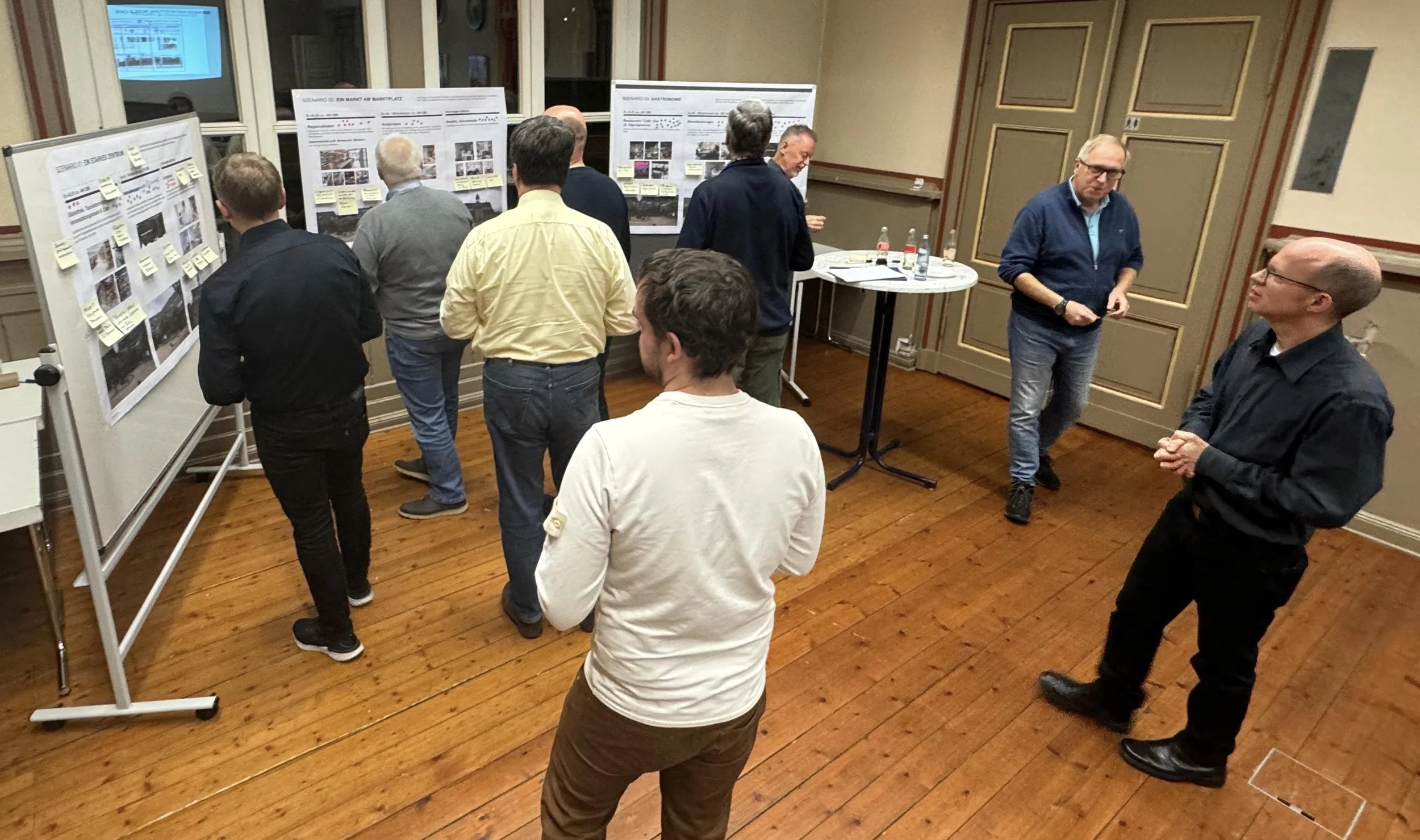 Foto vom Workshop im Projekt Aufwertung Marktplatz Vienenburg. Es sind mehrere Personen zu sehen, die rund um Whiteboards stehen, an denen Poster mit Bildern und Postits hängen.