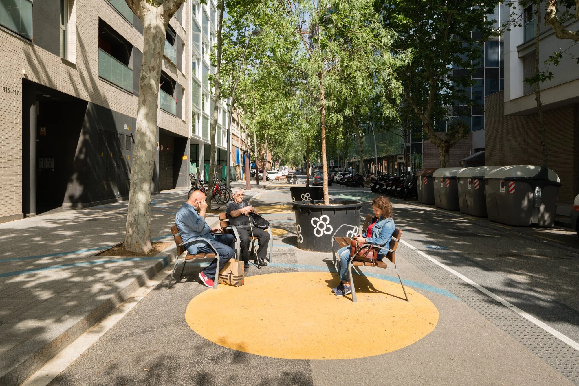 Drei Menschen sitzen auf Parkbänken in einer urbanen Straße mit Bäumen, Gebäuden und Müllcontainern, die Schatten spenden.