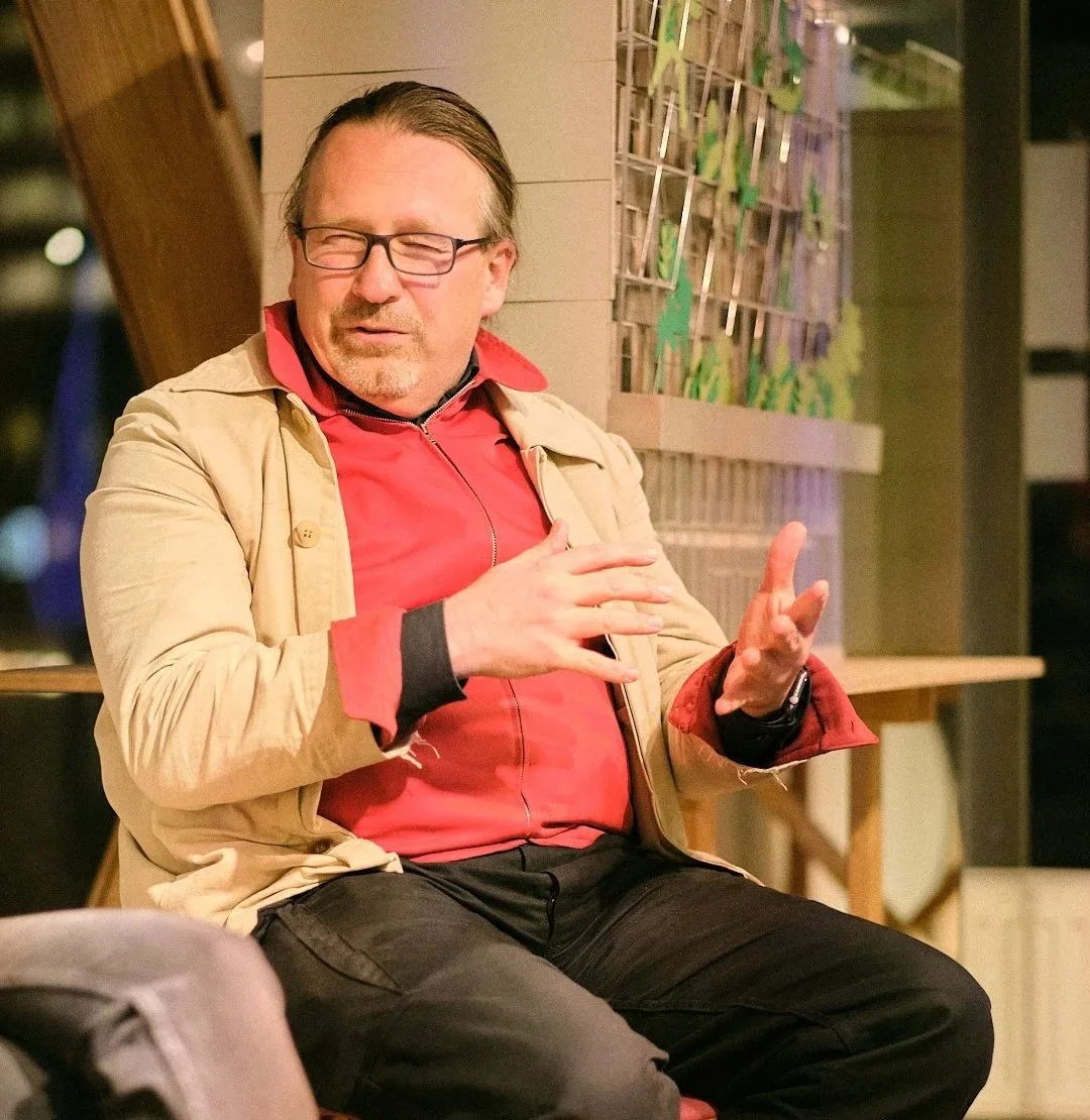 Ein Mann mit Brille und grauem Haar, in einem beige Mantel und roter Jacke, sitzt in einem Raum mit Holzelementen im Hintergrund, scheinbar im Gespräch.