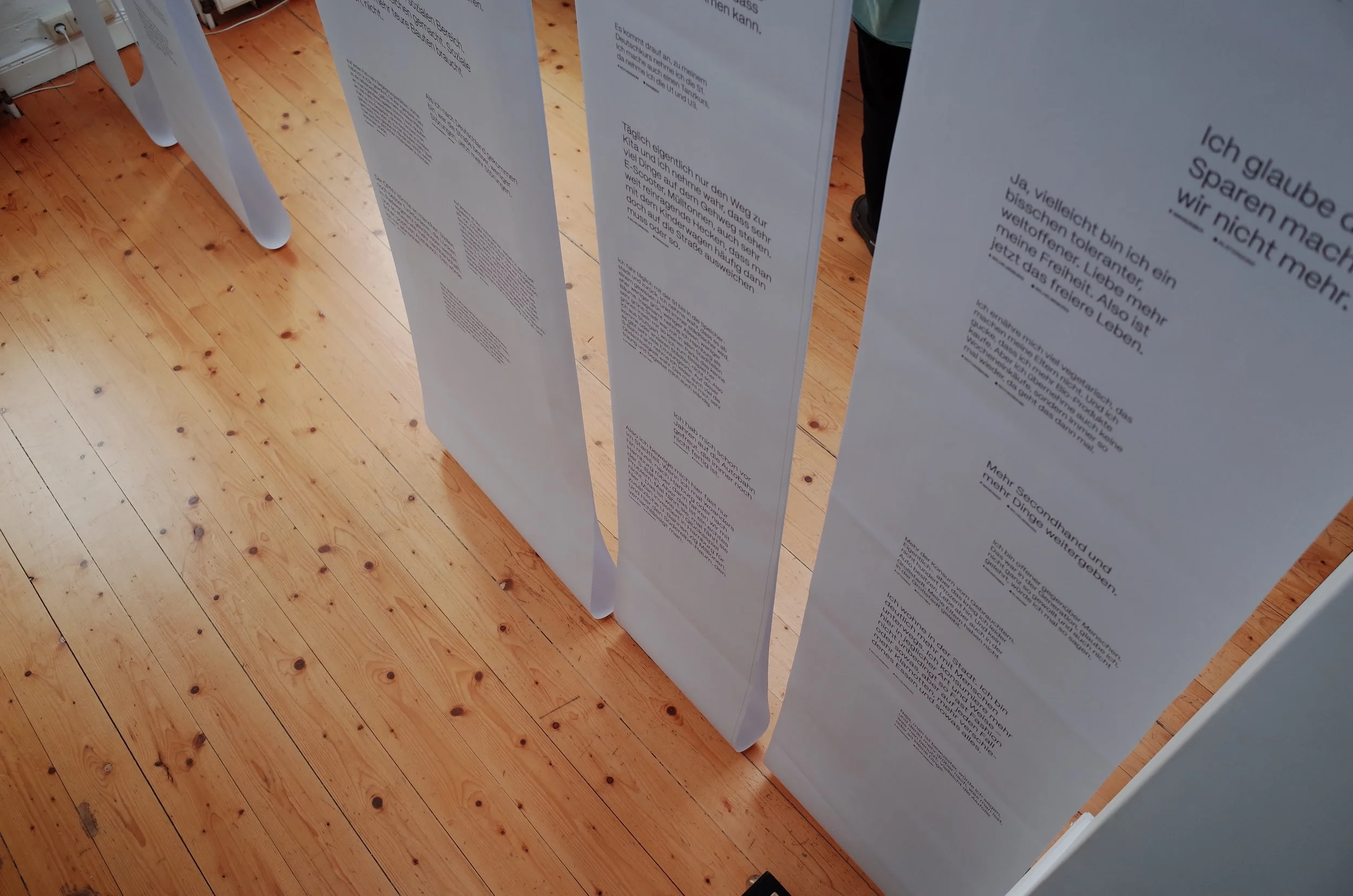 Ausstellungsraum im loft141 mit Bannern, die von der Decke hängen. Darauf Fragen und Antworten der Umfrage. Sustainability Week Hamburg 2026. Quartiersentwicklung. Beteiligung.