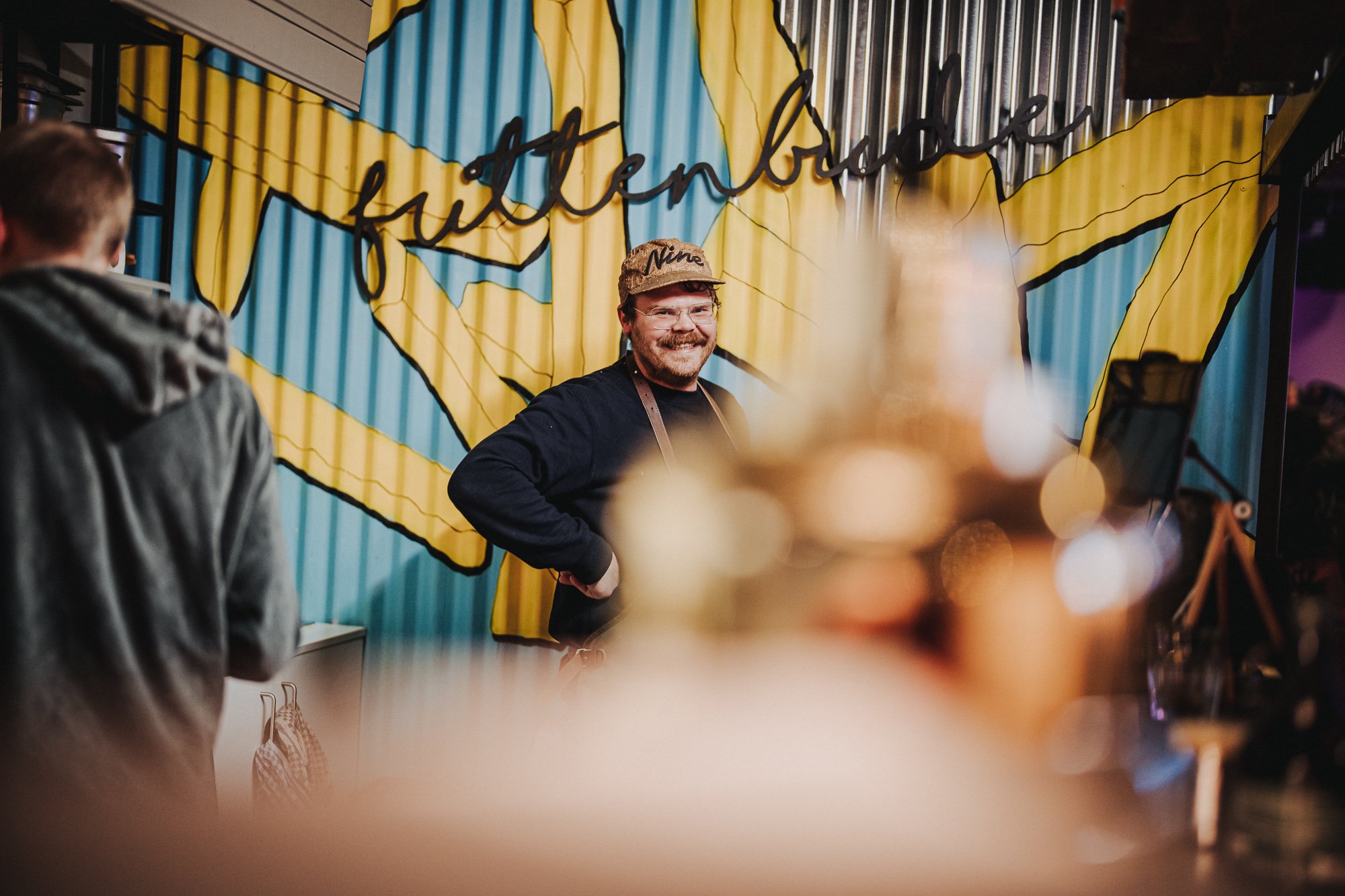 Ein lachender Mann mit Brille, schwarzem Pullover und Mütze steht vor einer Wand mit einer riesigen Deutschlandflagge, auf der das Wort "freiheit" geschrieben steht. Im Vordergrund sind unscharf Dekorationen.