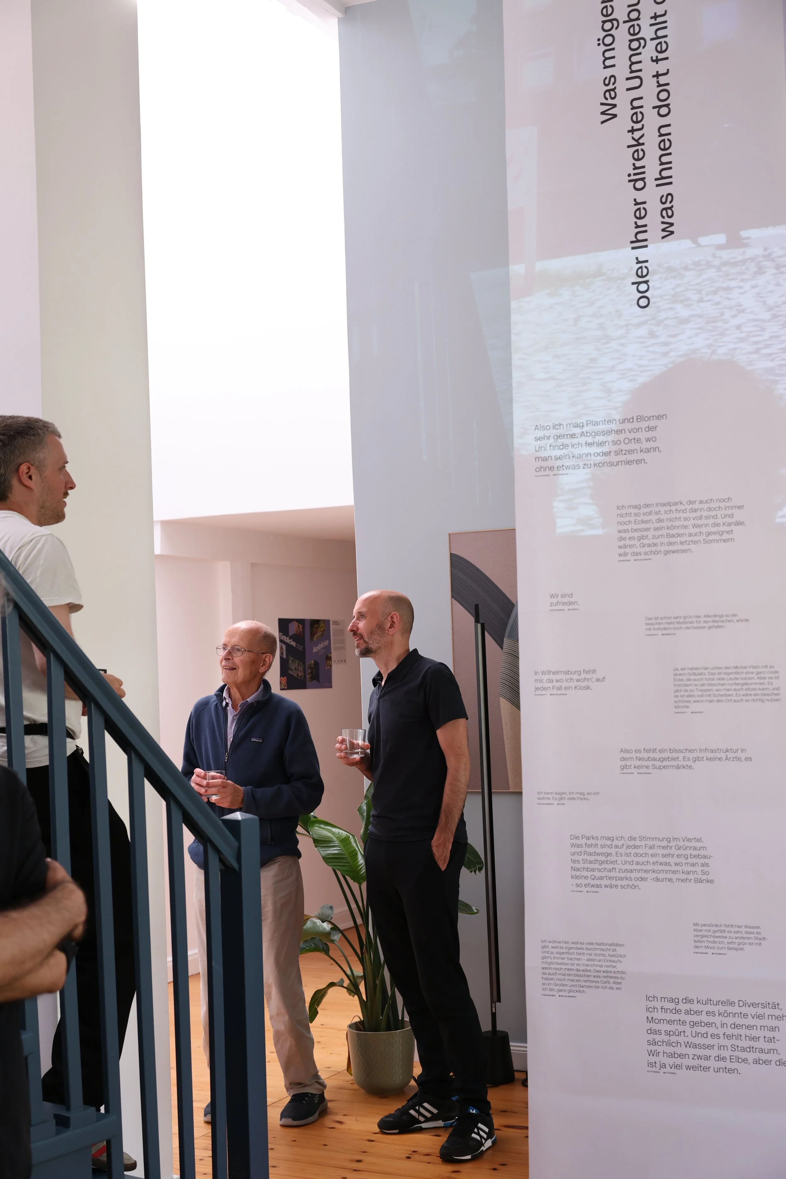 Drei Männer stehen in einem Gespräch in einem Raum mit weißen Wänden und großen Texttafeln, die von der Decke häng. Die Männer lächeln und halten Getränke. 
Hamburg Sustainability Week 2026. Ausstellung im Loft141.