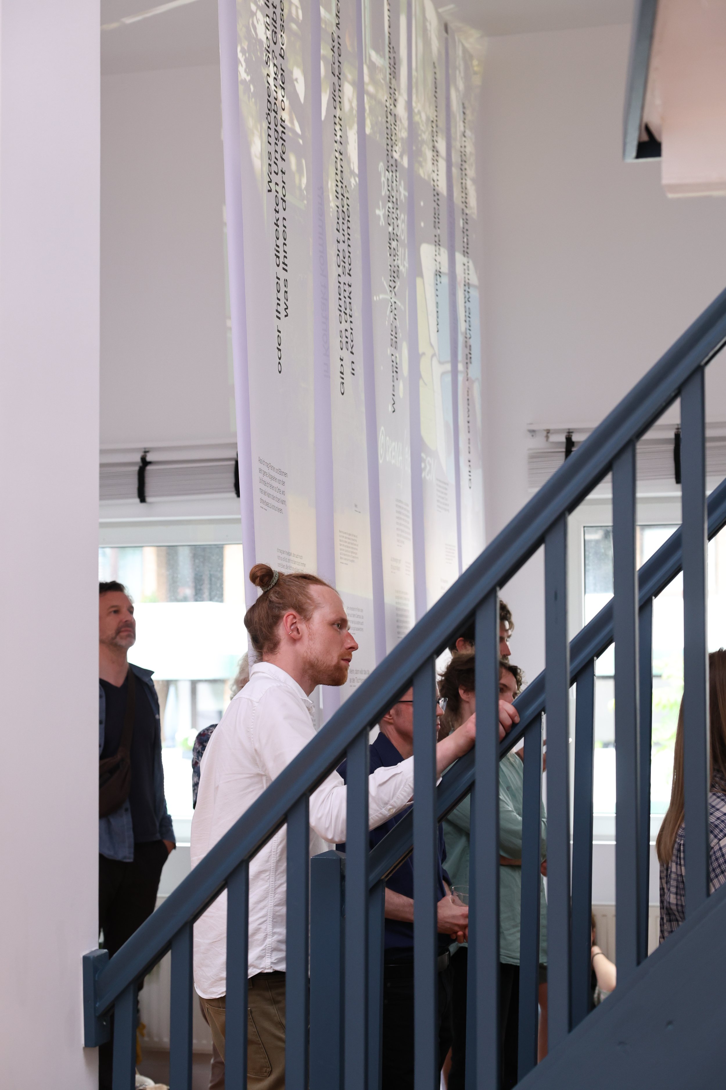Menschen in einem Ausstellungsraum diskutieren. Es gibt ein großes Banner mit Text von Decke, die Sonne scheint durch die Fenster. Eine Treppe ist im Vordergrund sichtbar.
Hamburg Sustainability Week 2026. Ausstellung im Loft141.