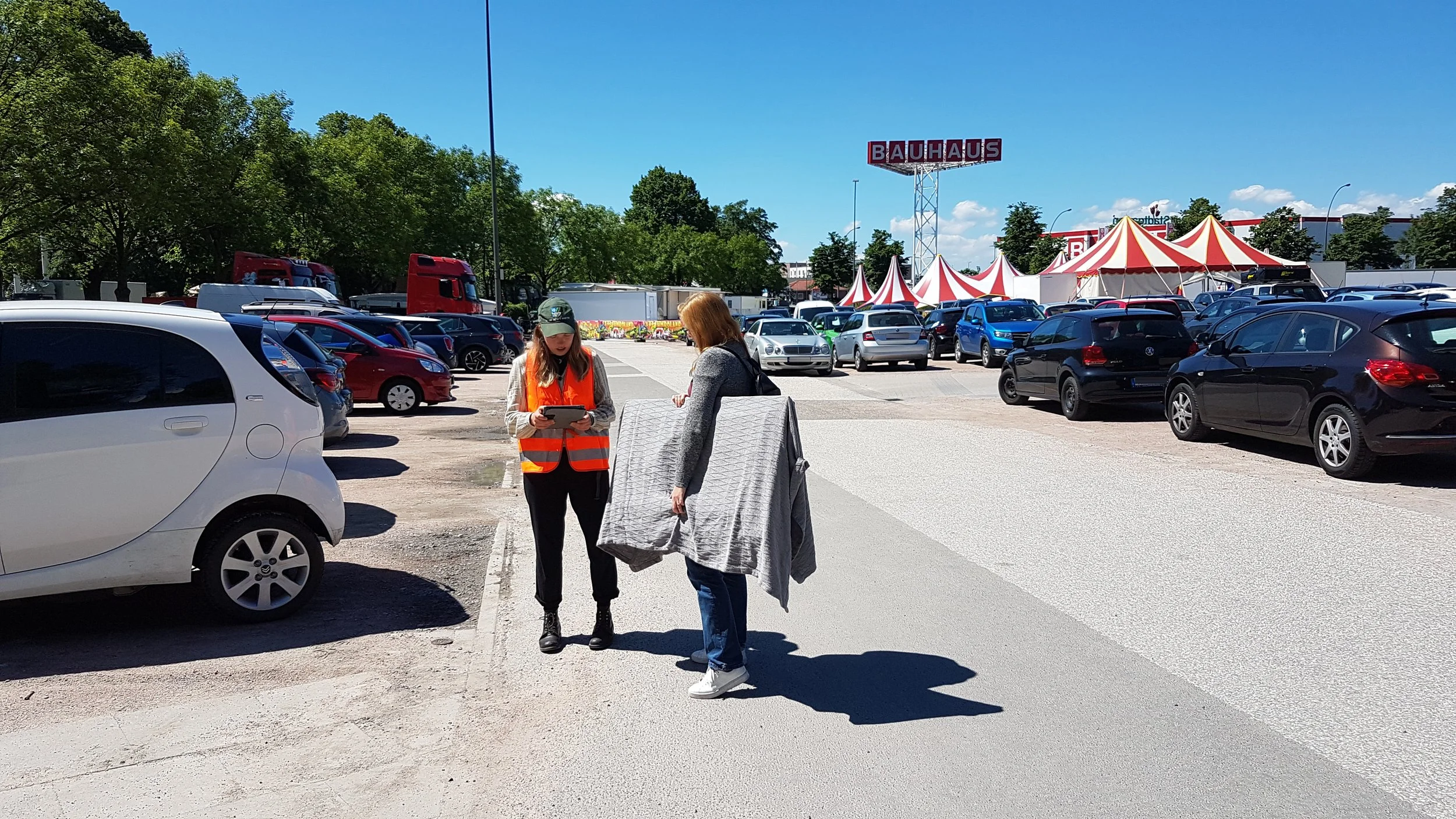 Frascatiplatz Hamburg Bergedorf. Zentral eine Frau mit Warnweste, die eine Besucherin des Platzes befragt. Drumherum parkende Autos.
