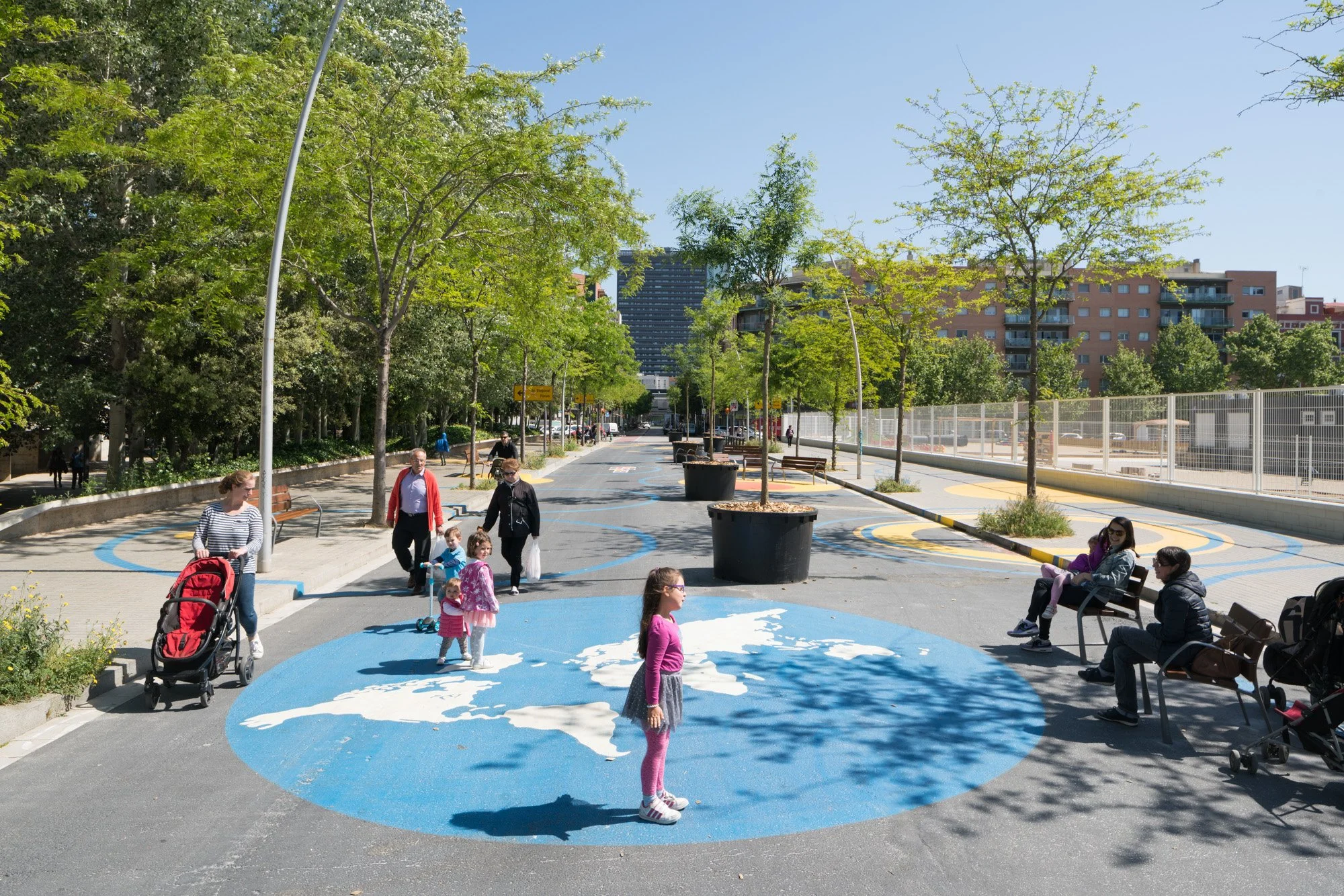 Ein Stadtpark mit Kindern, Erwachsenen und Sitzbänken, Bäumen, blauer Himmel und Hochhäuser im Hintergrund, mit künstlerischen Bodenmalereien auf dem Gehweg.
