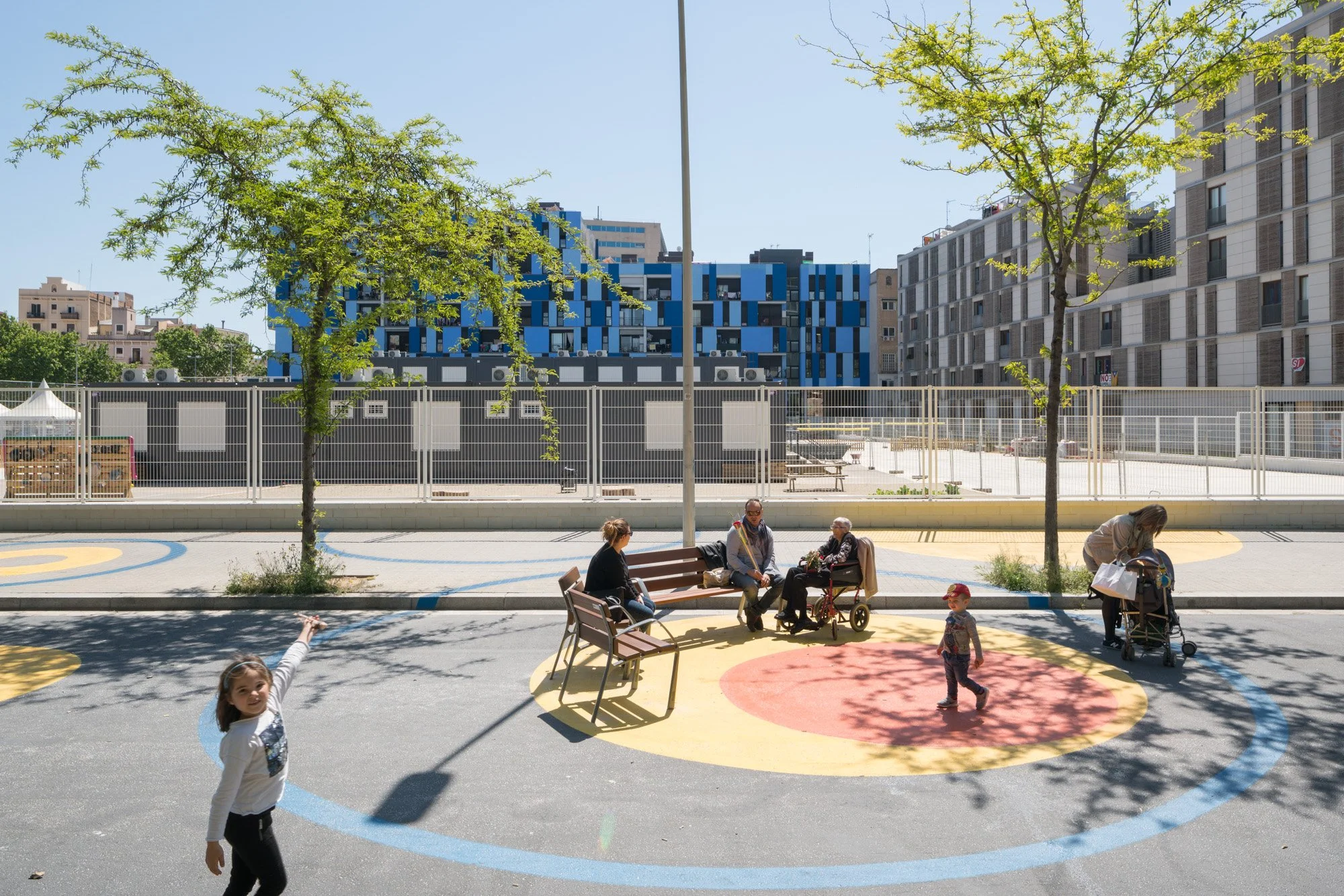 Kinder spielen auf einem Spielplatz im Freien, umgeben von Bäumen und modernen Gebäuden, bei sonnigem Wetter.