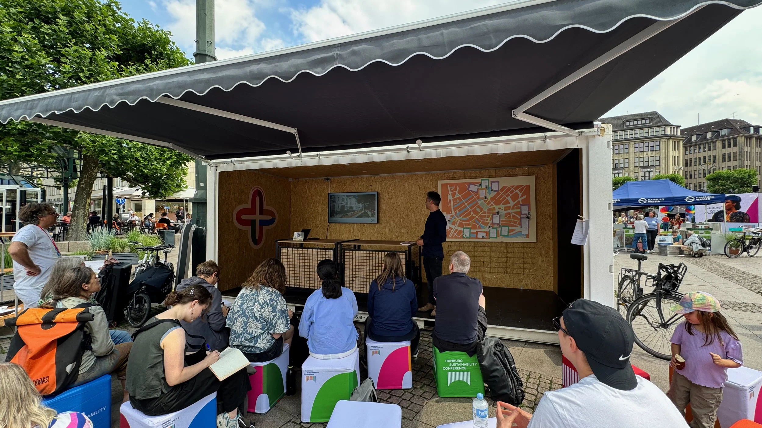 Lars Zimmermann von CITIES FOR FUTURE bei einem Vortrag in einem Container als Bühne, umgeben von zuhörenden Menschen. Hintergrund: Bäume, städtischer Architektur.
Hamburg Sustainability Week 2026, Zukunftsgarten auf dem Rathausmarkt Hamburg.