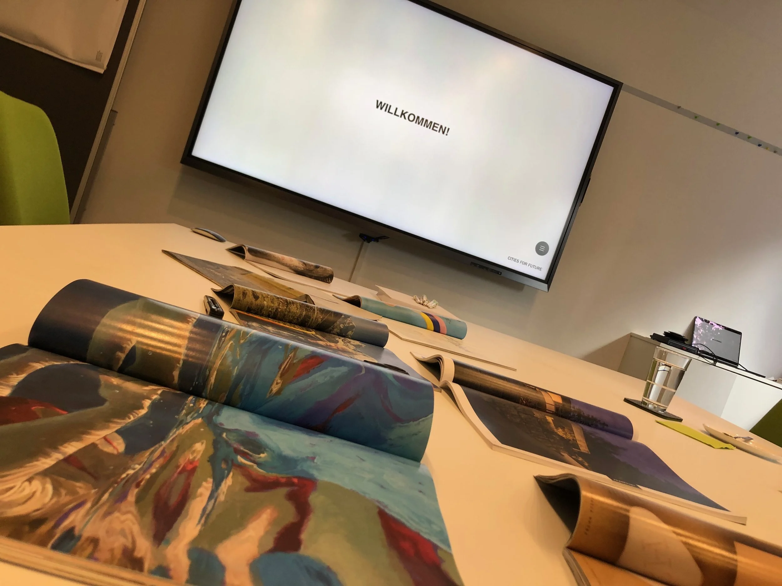 Foto eines worksops im Projekt Vialife Campus Bardenberg. Schiefer Blick in einen Konferenzraum ausgehend von einem Tisch mit aufgeklappten Magazinen. Am Bildschirm im Hintergrund steht "Willkommen".