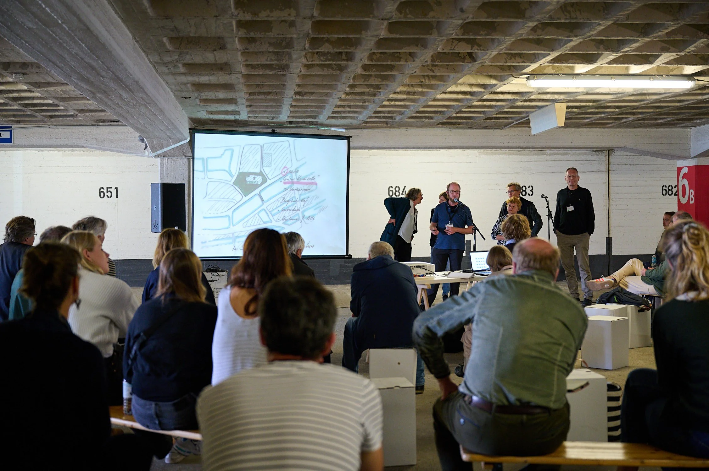 Projektbesprechung Alstadtküste Hamburg. Vorne Publikum, hinten mehrere Personen neben weißer Leinwand.