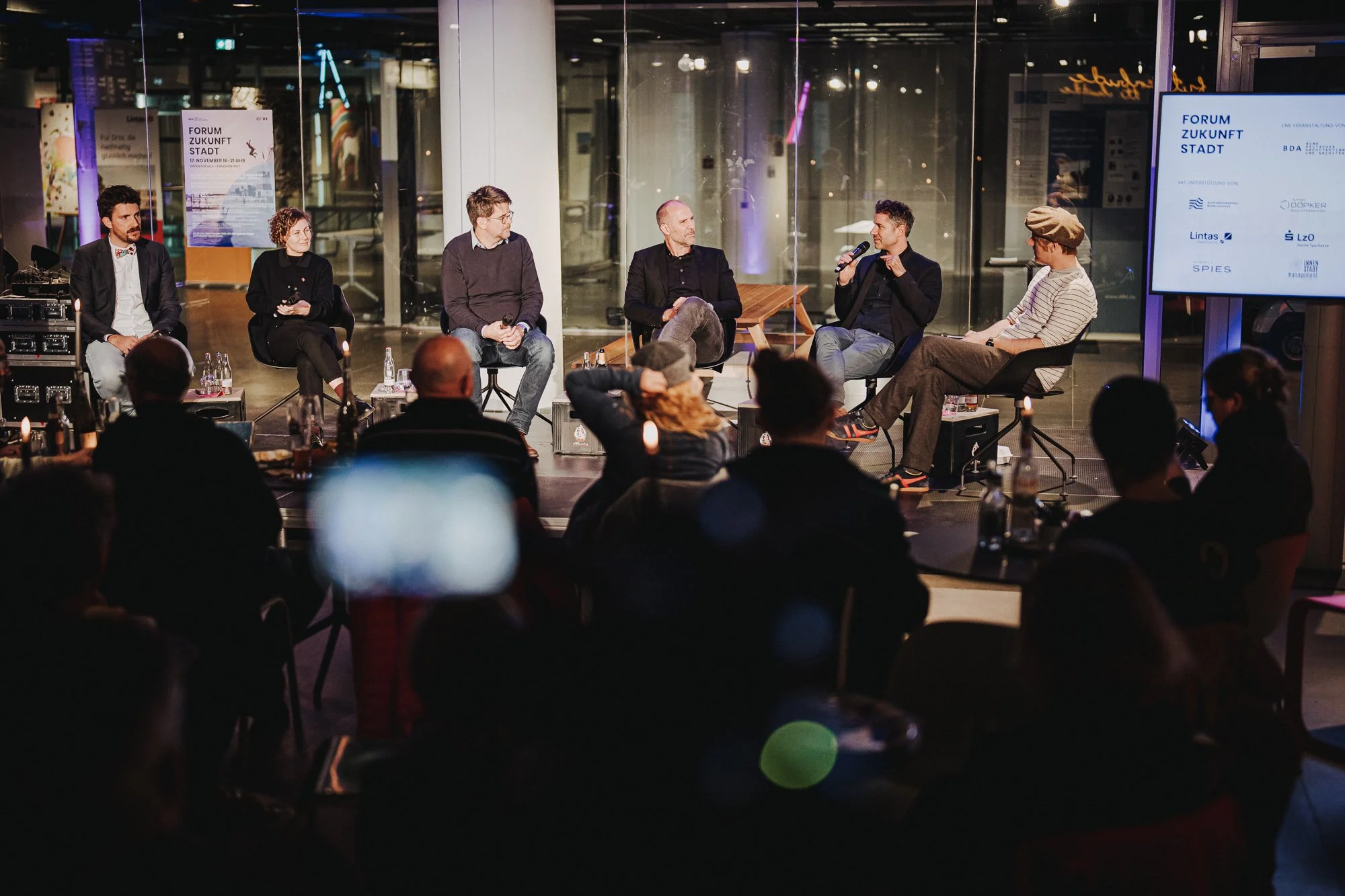 Eine Paneldiskussion bei einem Forum mit mehreren Sprechern auf der Bühne, im Hintergrund ein Bildschirm und Publikum im Vordergrund.