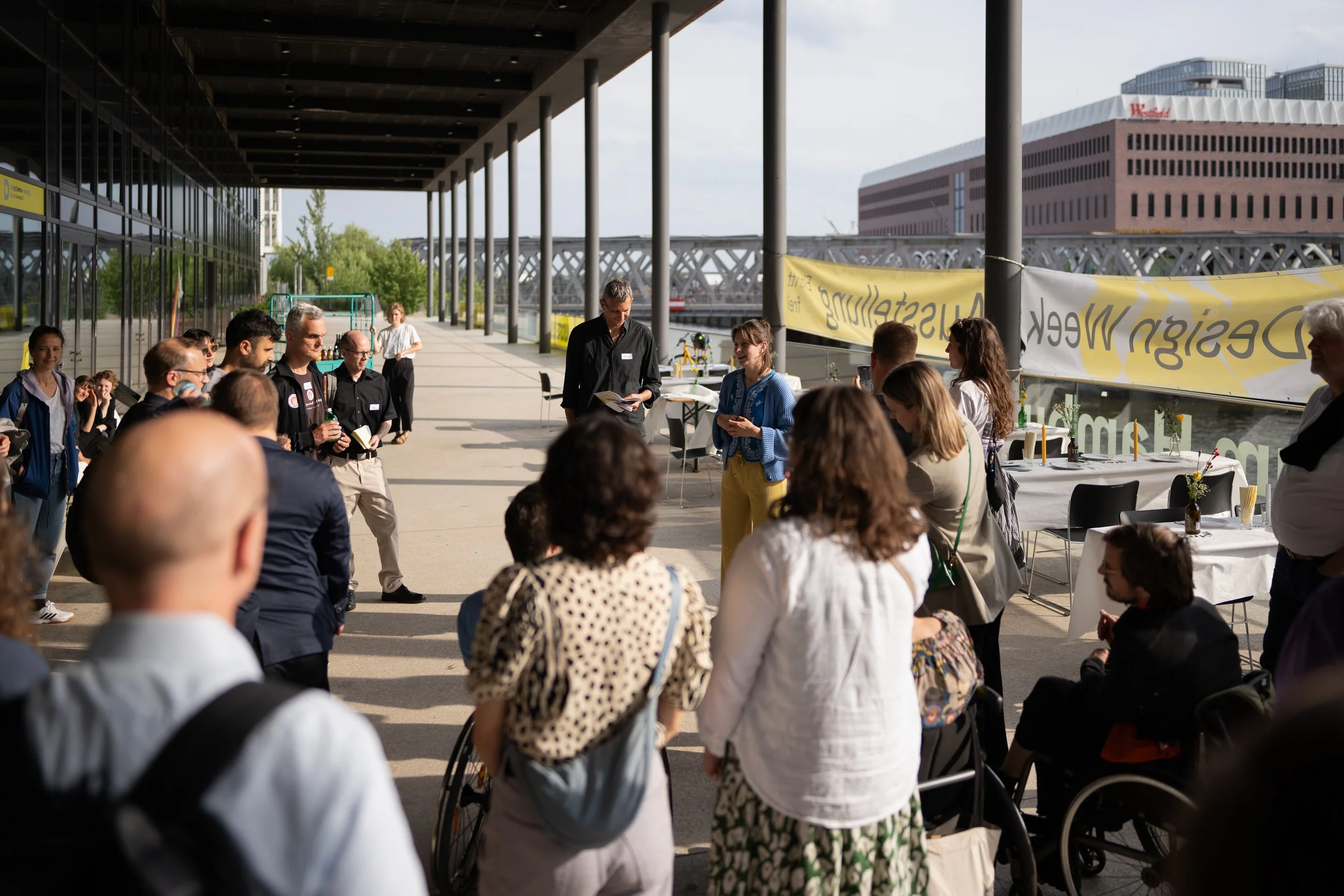 Gruppen von Menschen bei einer Veranstaltung im Freien unter einem überdachten Bereich, einige sitzen, andere stehen, während eine Organisation oder Diskussion stattfindet.