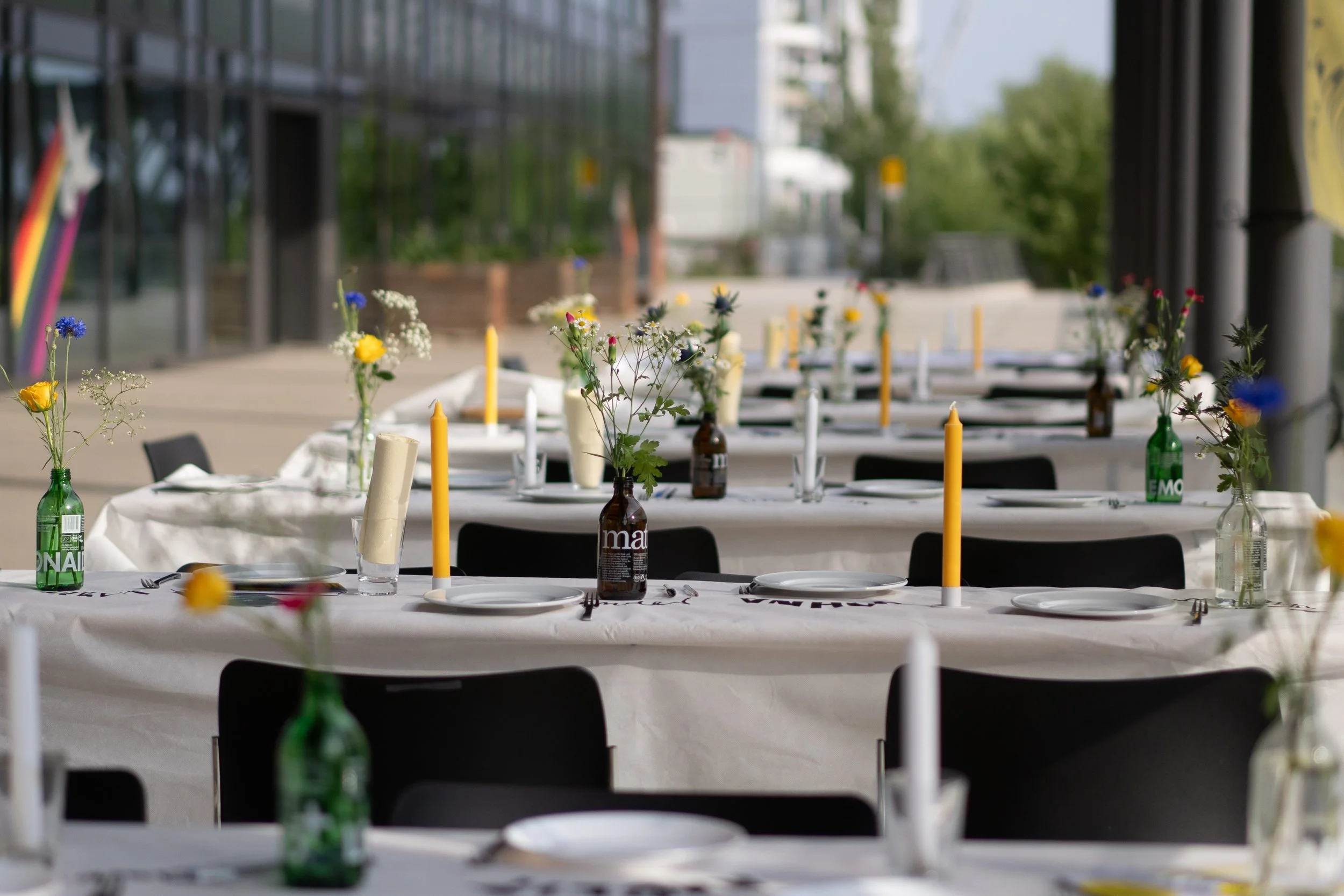 Ein festlich gedeckter Tisch im Außenbereich mit weißen Tischtüchern, kleinen Blumenvasen, gelben Kerzen und schwarzen Stühlen, bereit für eine Veranstaltung.