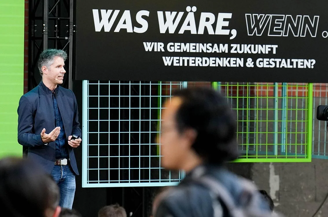Lars Zimmermann von CITIES FOR FUTURE hält einen Vortrag vor Publikum vor einem großen Bildschirm mit deutschem Text, der Fragen zur Zukunft stellt während der #ZNV23.
