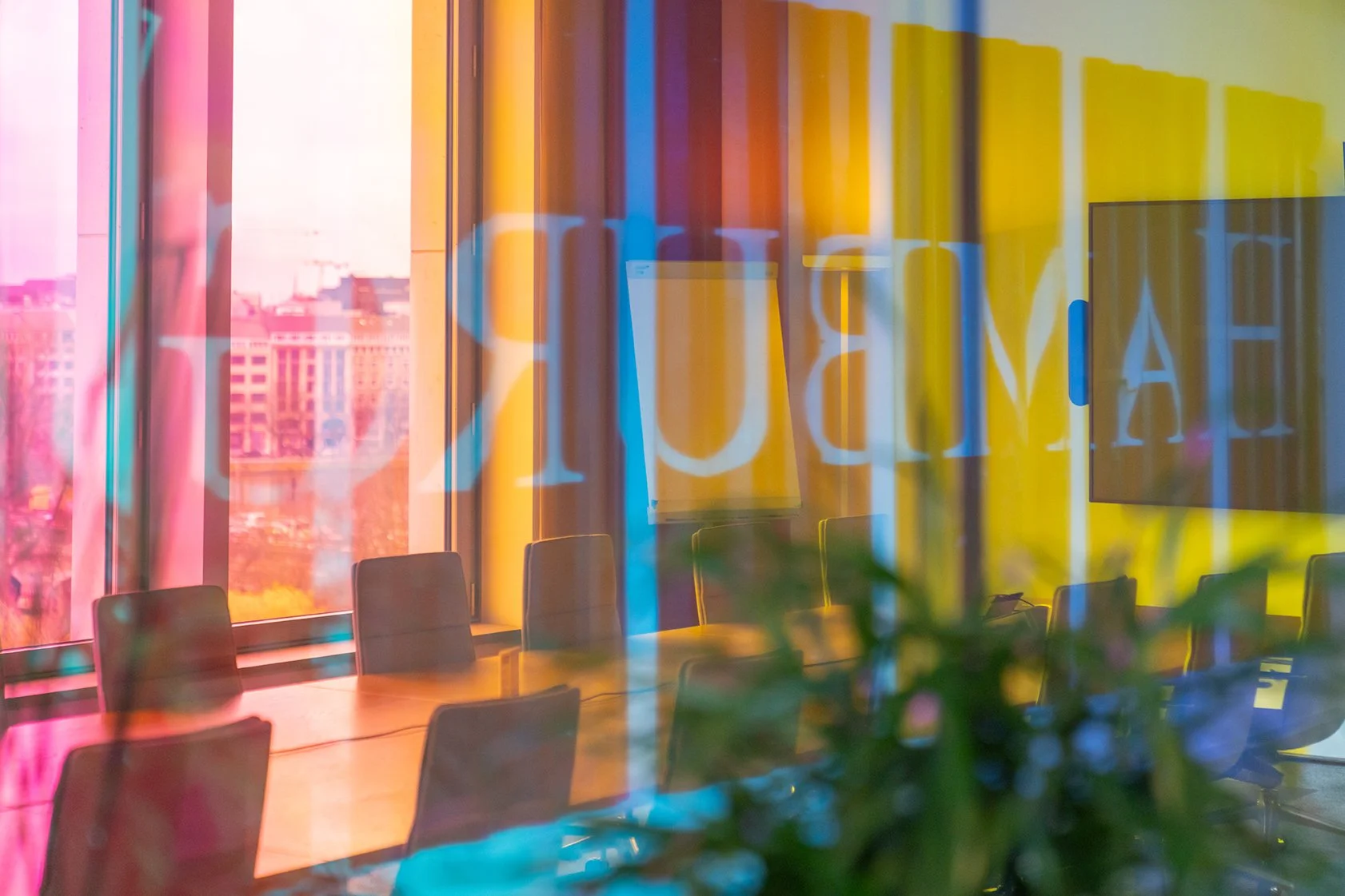 Dentsu Office:  Blick durch bunte Galsscheiben. Dahinter ein Konferenzraum mit vielen Stühlen an einem langen Tisch. Themenbereich: Interiorgestaltung, Innenarchitektur, Placemaking.