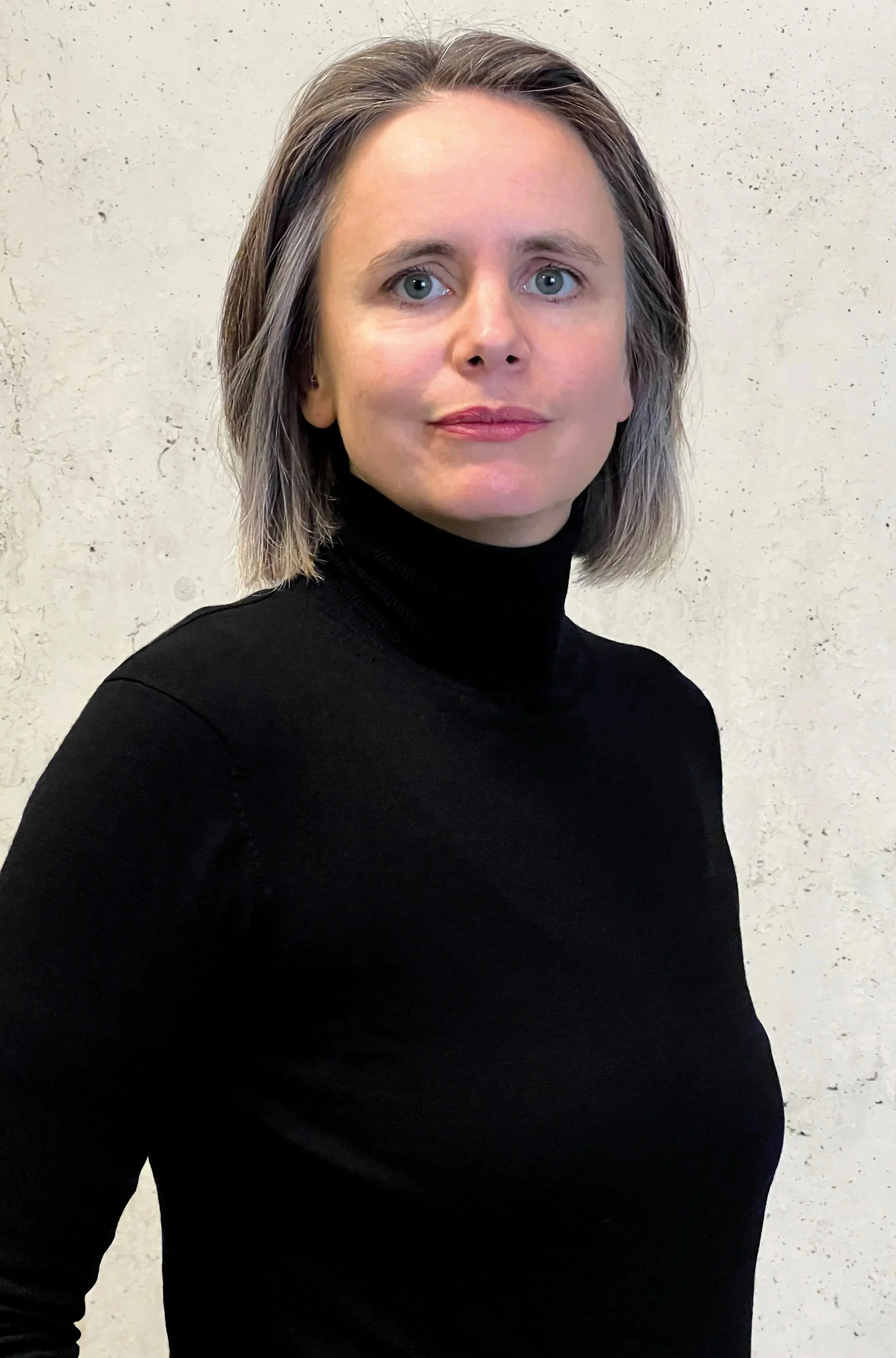 Julia Miosga mit schulterlangem grauem Haar, trägt schwarzen Rollkragenpullover, steht vor einer hellen Wand, direkter Blick in die Kamera. Julia Miosga ist Digital Landschafts Gärtnerin.