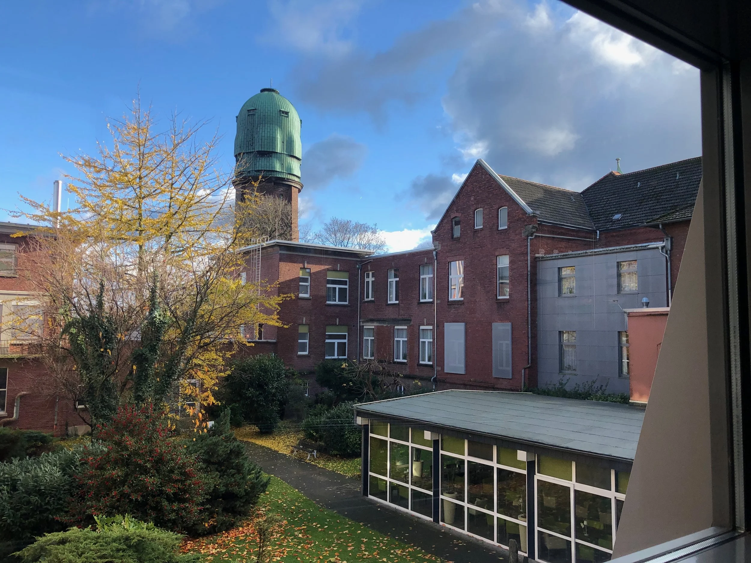 Blick durch ein Gebäudefenster auf den Vialife Campus Bardenberg_Blick auf Gebäudeteile des Arelas in rotem Backstein und mit Wasserturm. Vorne Sprossenfensterfront.