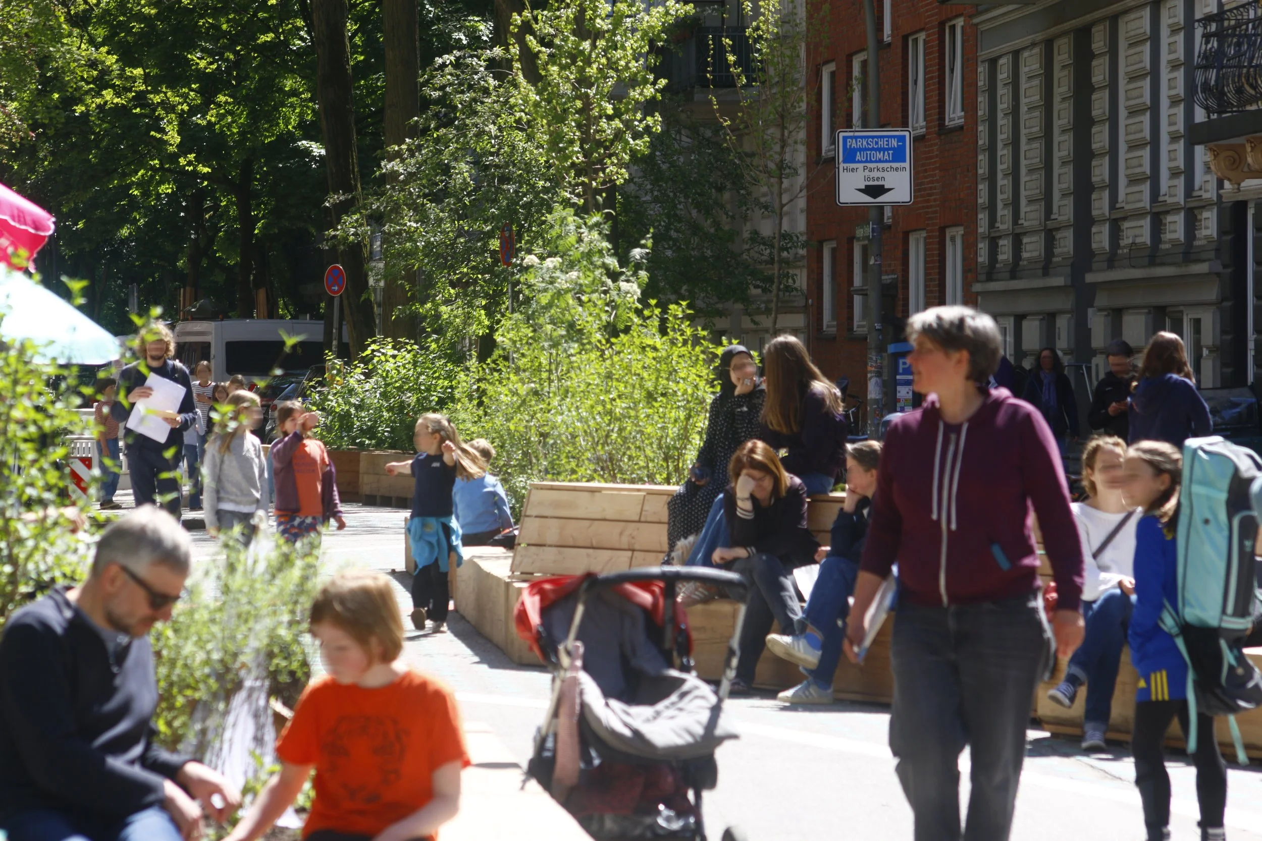 1. Hamburger Schulstraße Rellinger Straße Hamburg Eimsbüttel, Aufnahme vom Eröffnungstag 8. Mai 2026. Viele Erwachsene und Kinder, spielend und sitzend auf Holzbänken. Viel Begrünung. Themenbereiche: Superblocks, Aufenthaltsqualität, Mobilität.
