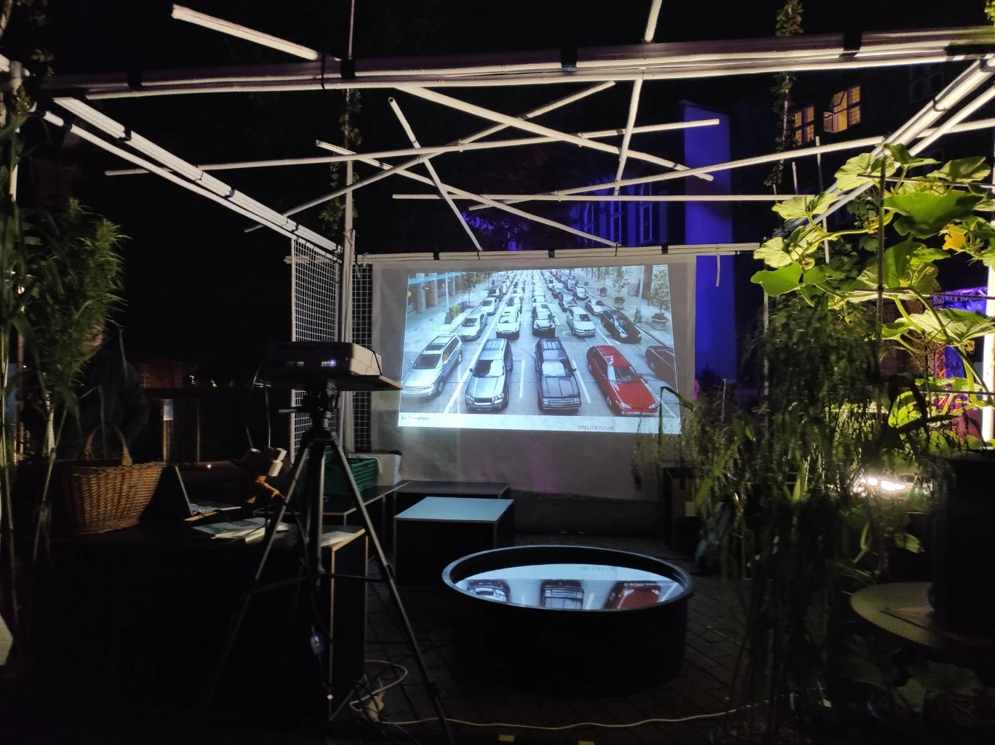 SAMMELSTELLE Pavillon auf dem Dach des Hansemuseums in Lübeck, in dem ein Nachtprojektor eine Straße mit Autos anzeigt, umgeben von Pflanzen, mit Dekoration und Kabeln.