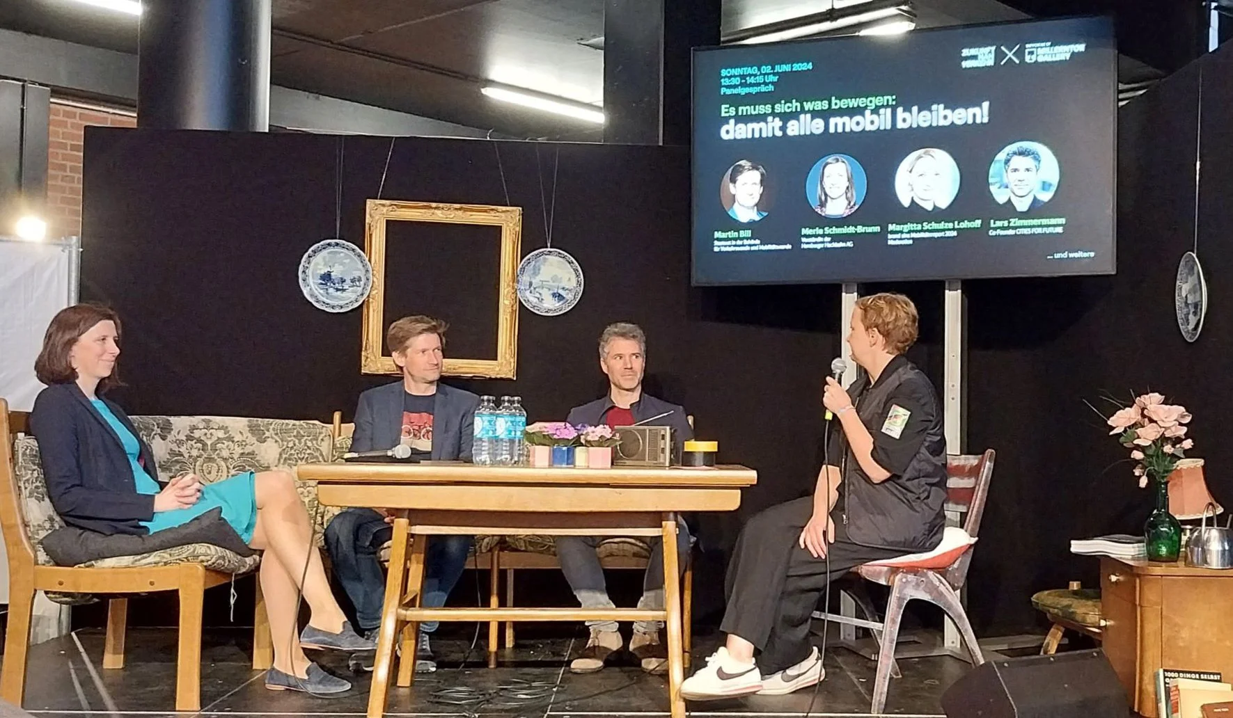 Menschen bei einer Podiumsdiskussion auf einer Bühne, mit einem großen Bildschirm im Hintergrund, der Informationen zur Veranstaltung zeigt. Martin Bill, Merle Schmidt-Brunn