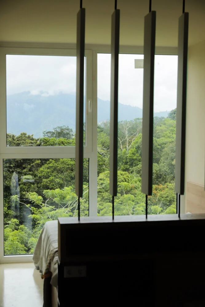 Ein Schlafzimmer mit großen Fenstern, die eine grüne, bewaldete Berglandschaft zeigen, und modernen vertikalen Leuchten oder dekorativen Elementen im Raum.