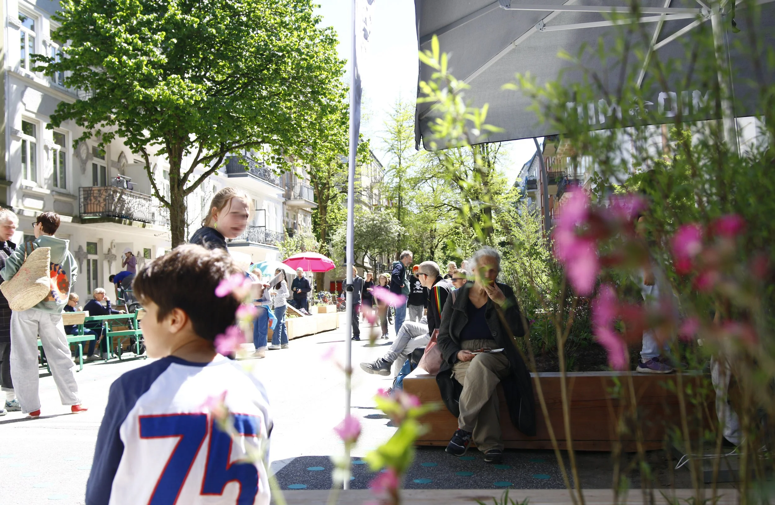 Schulstraße Rellinger Straße, 8. Mai 2026. Viele Erwachsene und Kinder bewegen sich. Viel Begrünung. Themenbereiche: Superblocks, Aufenthaltsqualität, Mobilität.