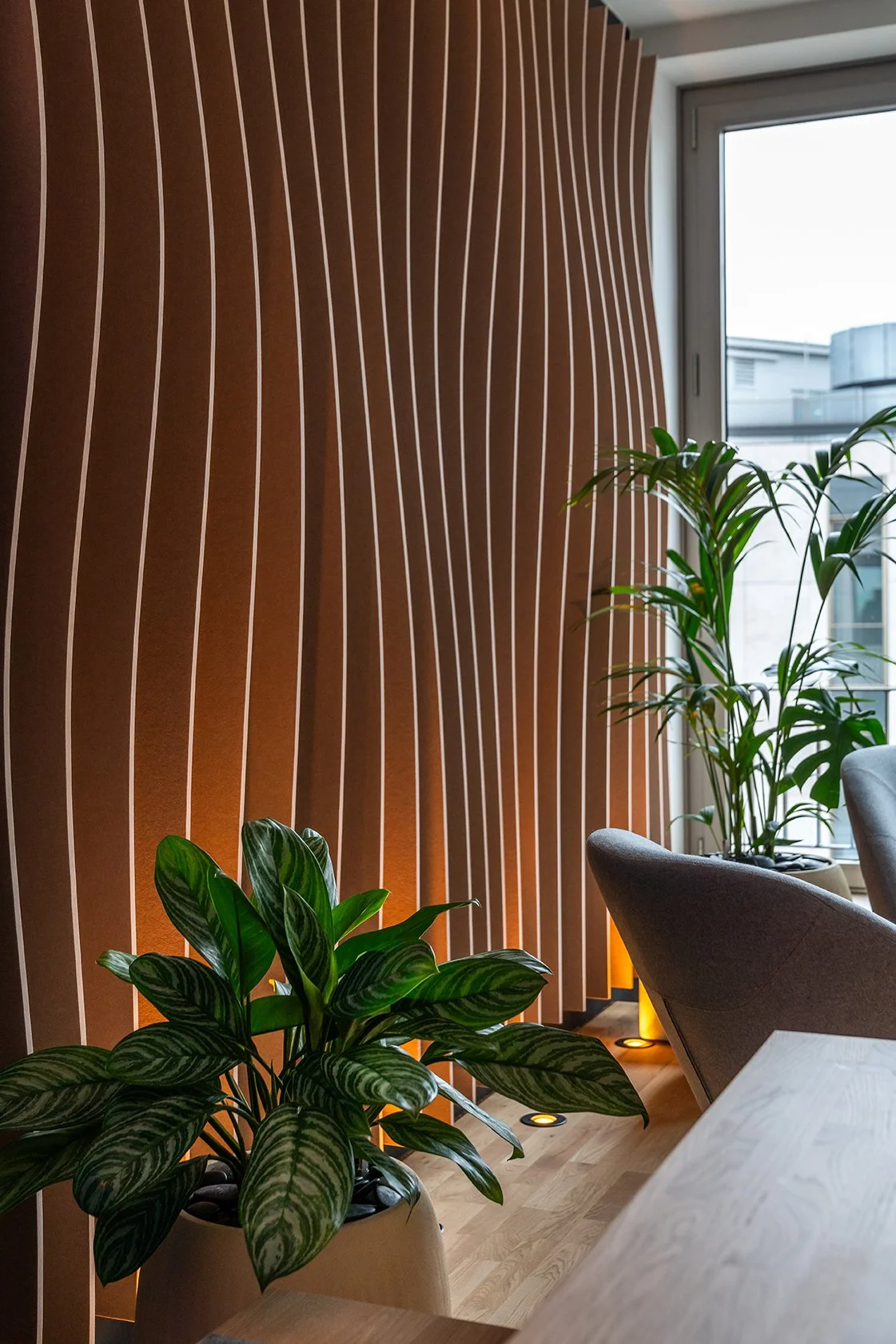 Dentsu Office: Raum mit Holzlamellenwand auf der linken Seite. Rechts Pflanzen, runde Stühle und ein Tisch. Hintergrund: Fensterblick. Themenbereich: Interiorgestaltung, Innenarchitektur, Placemaking.