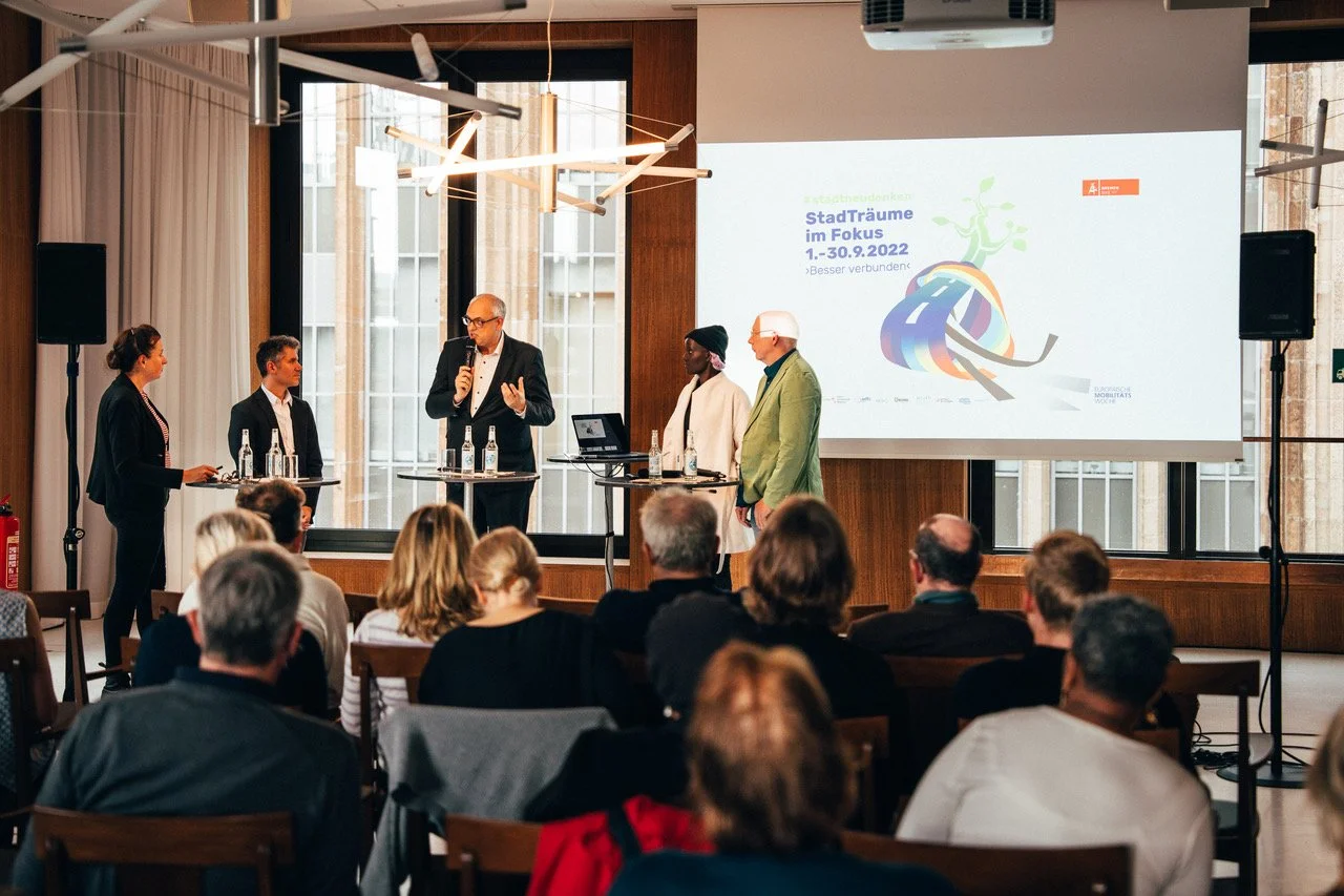 StadtTraum Bremen 2022. Menschen bei einem Vortrag oder Paneldiskussion in einem Konferenzraum, vorne eine Gruppe von fünf Sprechern, einer spricht gerade, im Hintergrund eine Leinwand mit Präsentation. U.a. Lars Zimmermann von CITIES FOR FUTURE