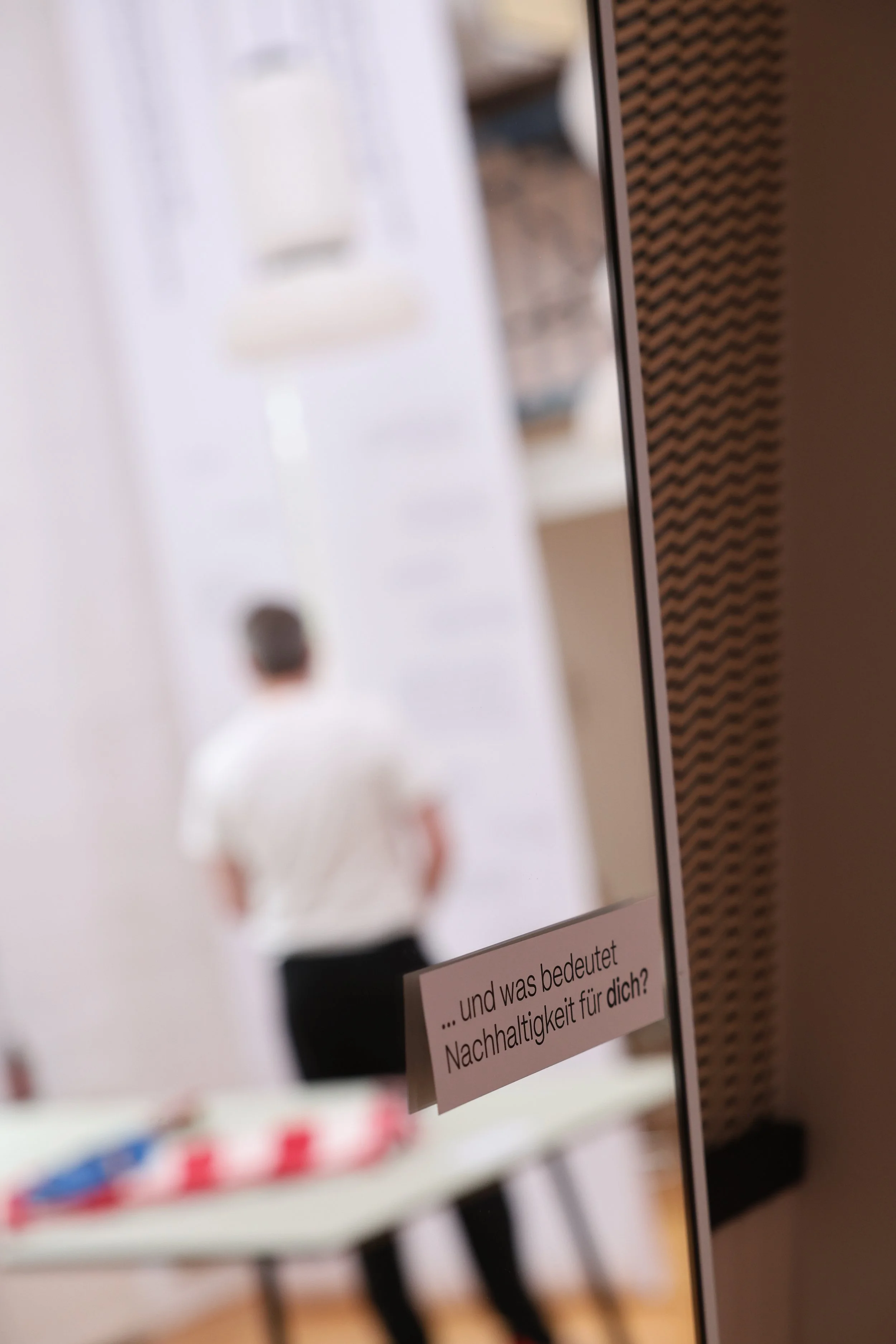 Nahaufnahme von Ausstellungsstücken auf einem Tisch zu Treffpunkten im Quartier, einer Frage der Umfrage. Im Hintergrund steht ein Mann vor den weißen Textbannern von der Decke.
Hamburg Sustainability Week 2026. Ausstellung im Loft141.