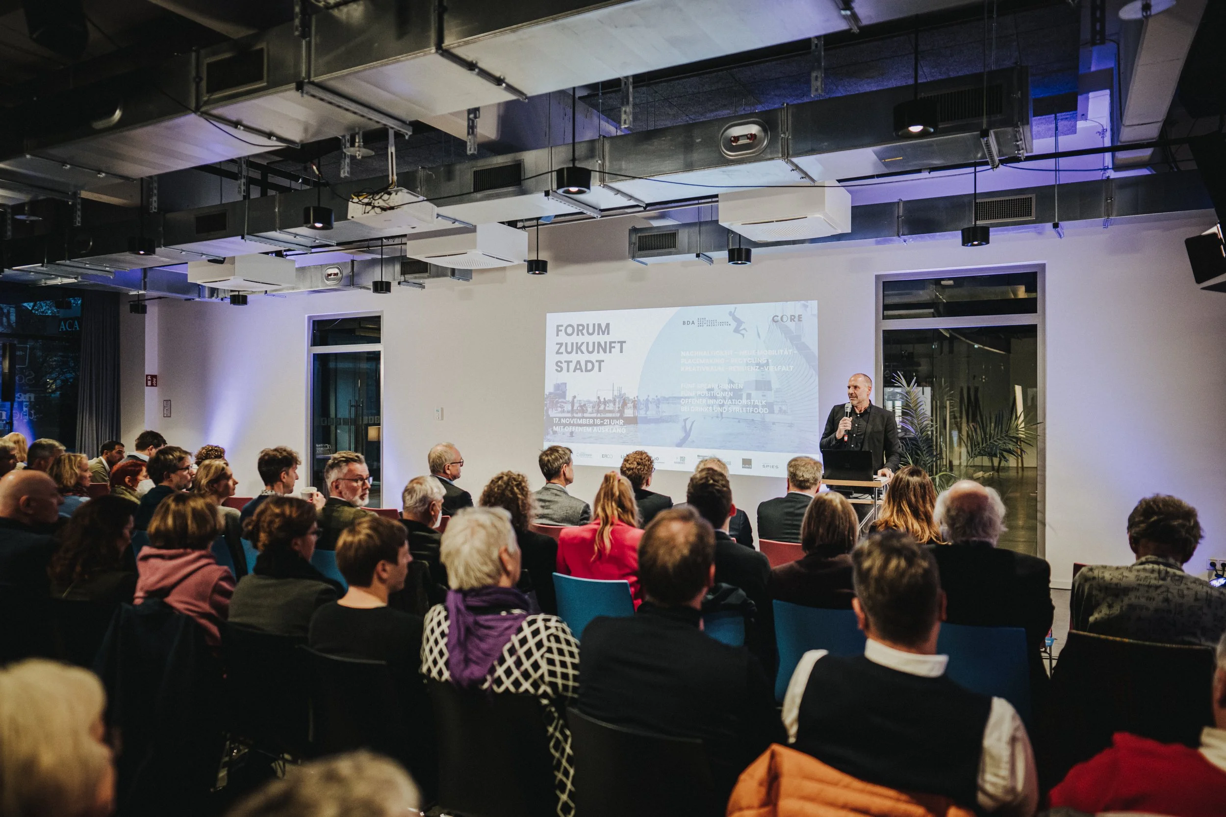 Ein Vortrag in einem modernen Veranstaltungsraum mit einem Publikum und einem Redner an einem Pult vor einer Projektionswand, die Informationen zum "Forum Zukunft Stadt" anzeigt.