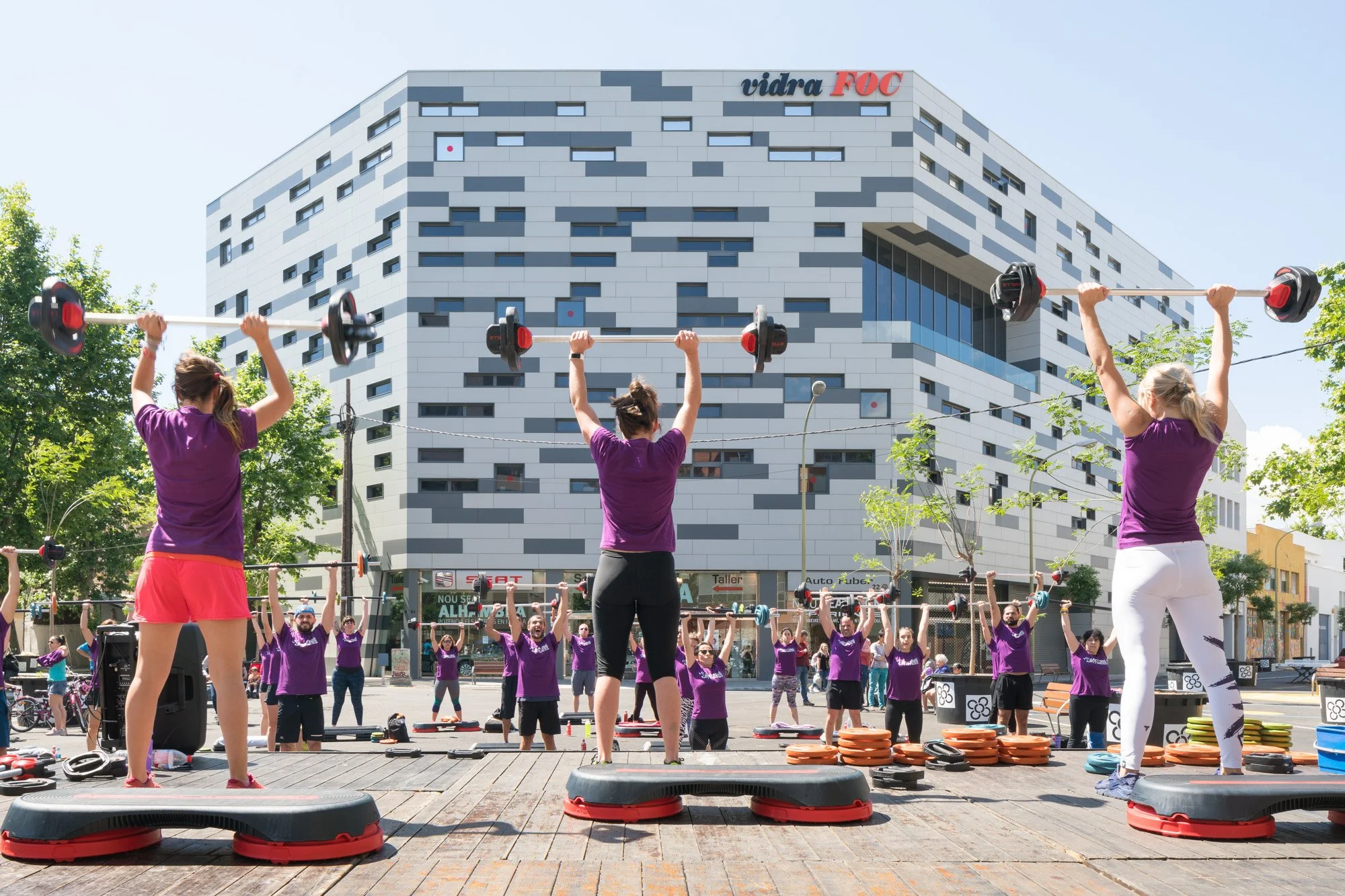 Gruppen von Menschen machen ein Outdoor-Fitness-Workout mit Gewichten und Steppern vor einem modernen Gebäude an einem sonnigen Tag.