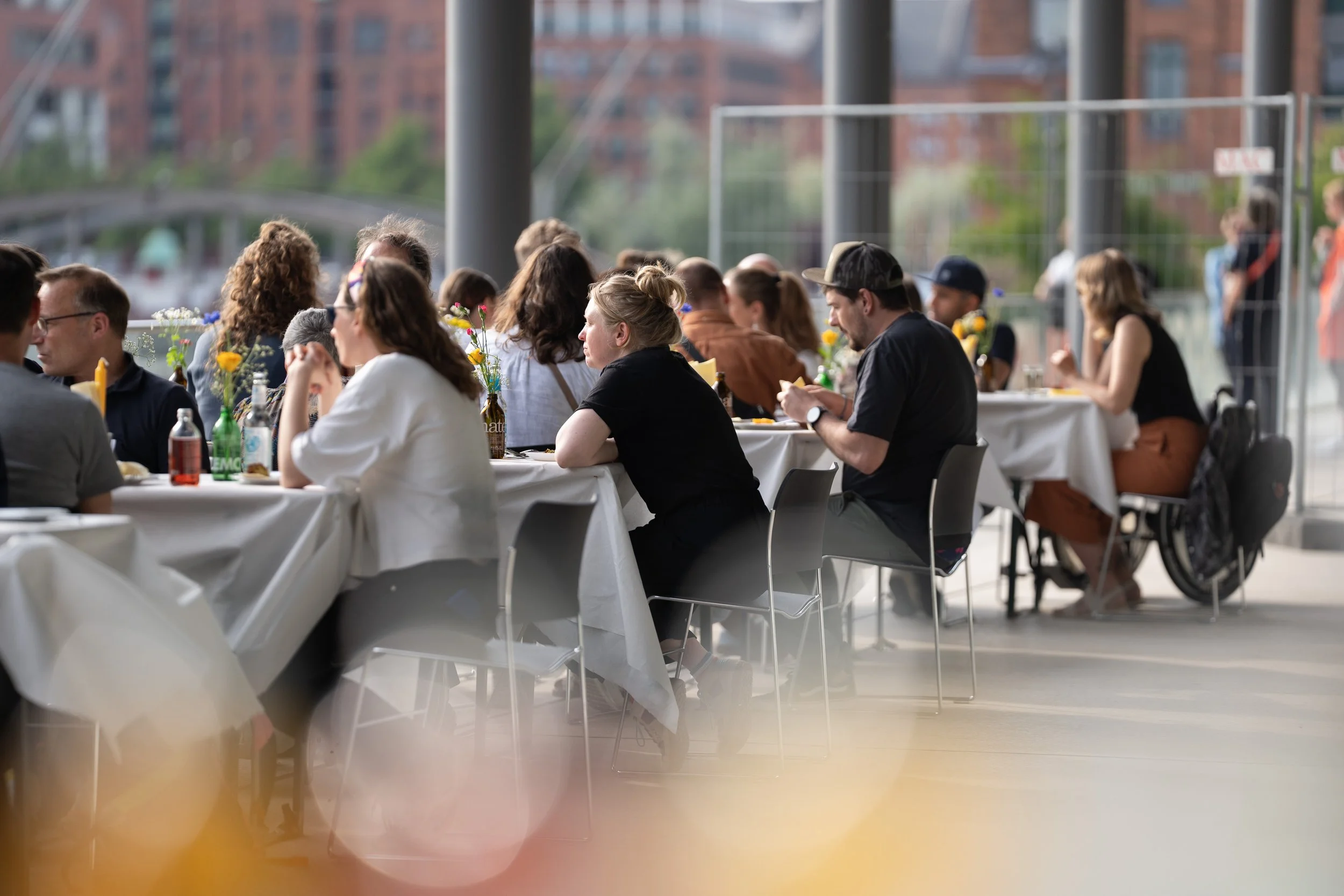 Menschen sitzen an langen Tischen bei einer Outdoor-Veranstaltung, umgeben von Blumenvasen, im Hintergrund sind Gebäude sichtbar.