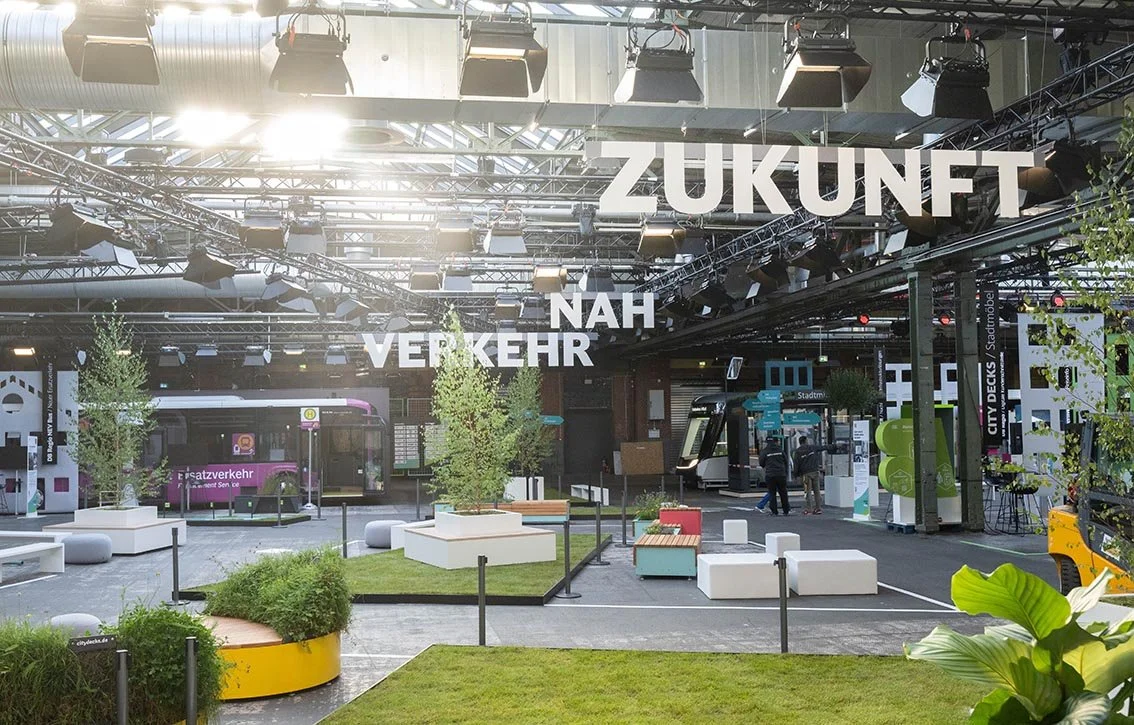 #ZNV23 Innenansicht einer modernen urbanen Haltestelle mit Bussen, Sitzgelegenheiten und Pflanzen, überdacht mit einem Stahlträgerdach und einer Beschilderung, die 'Zukunft Nahverkehr' zeigt.
