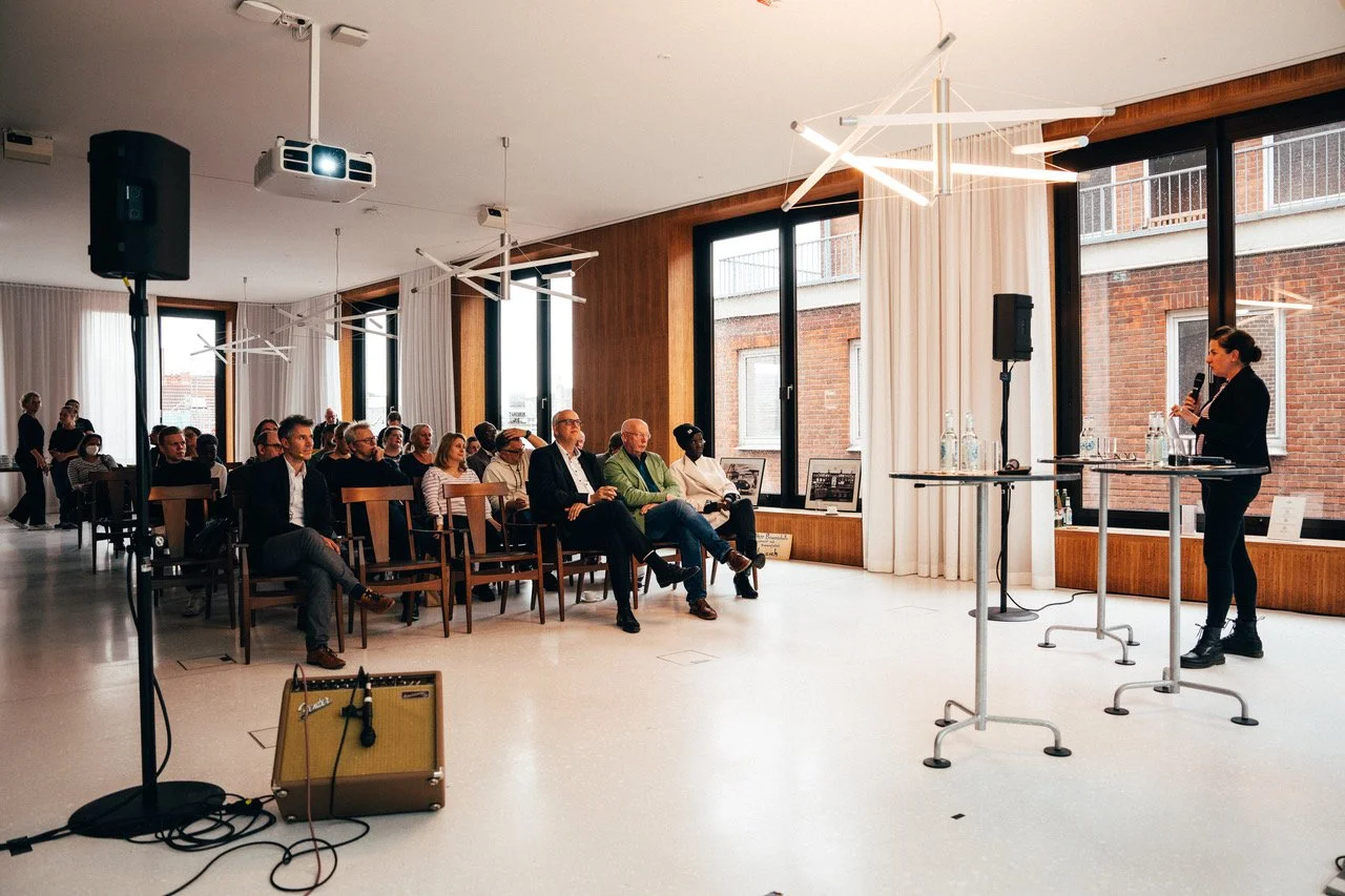Frau hält eine Rede vor einem Publikum in einem hellen Raum mit großen Fenstern und moderner Beleuchtung.