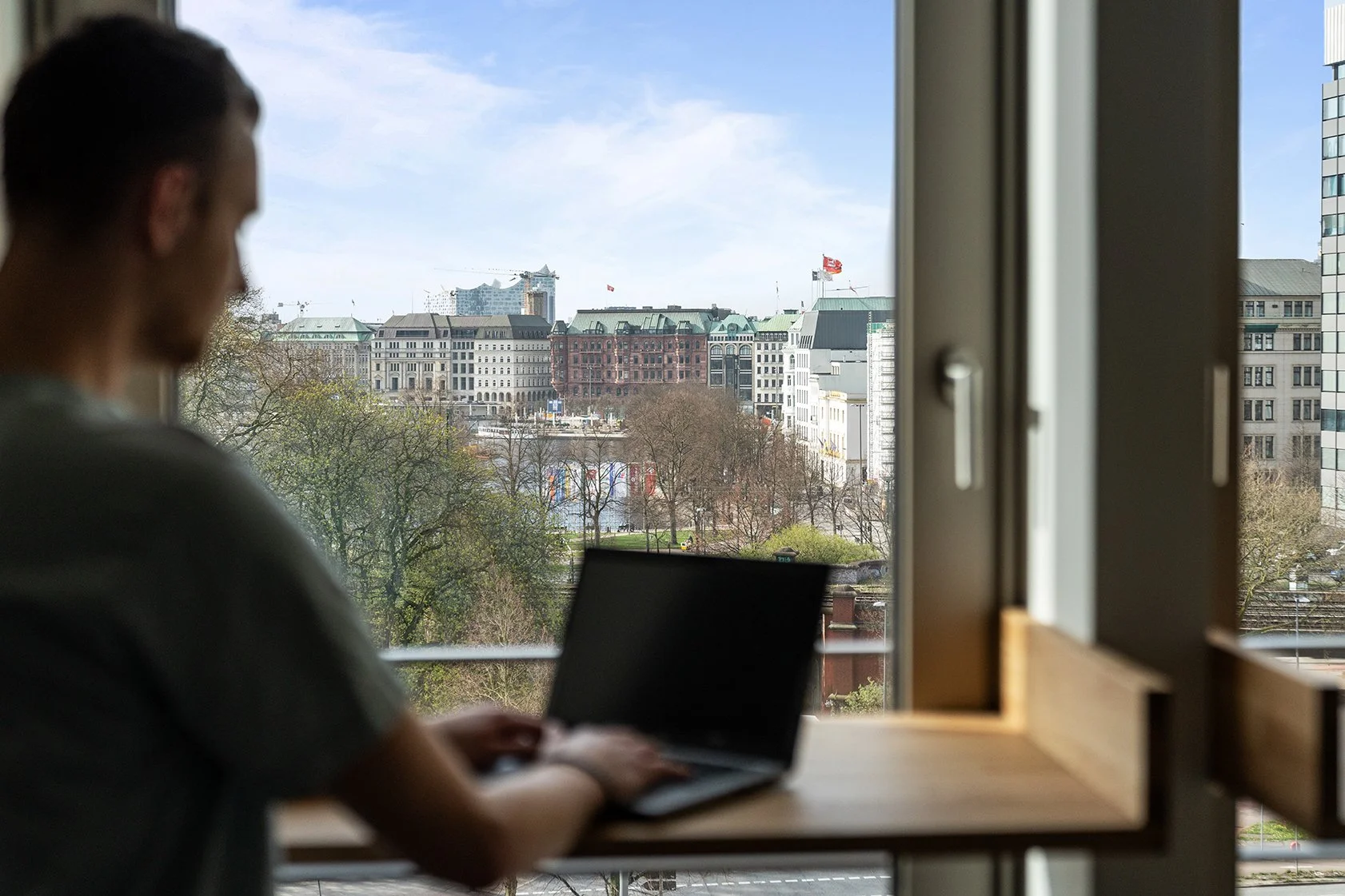 Dentsu Office: Vorne ist unscharf eine Person an einem Holztisch mit Laptob zu sehen. Hintergrund Blick durch Fenster auf die Alster und umliegende Gebäude in Hamburg.Themenbereich: Interiorgestaltung, Innenarchitektur, Placemaking.
