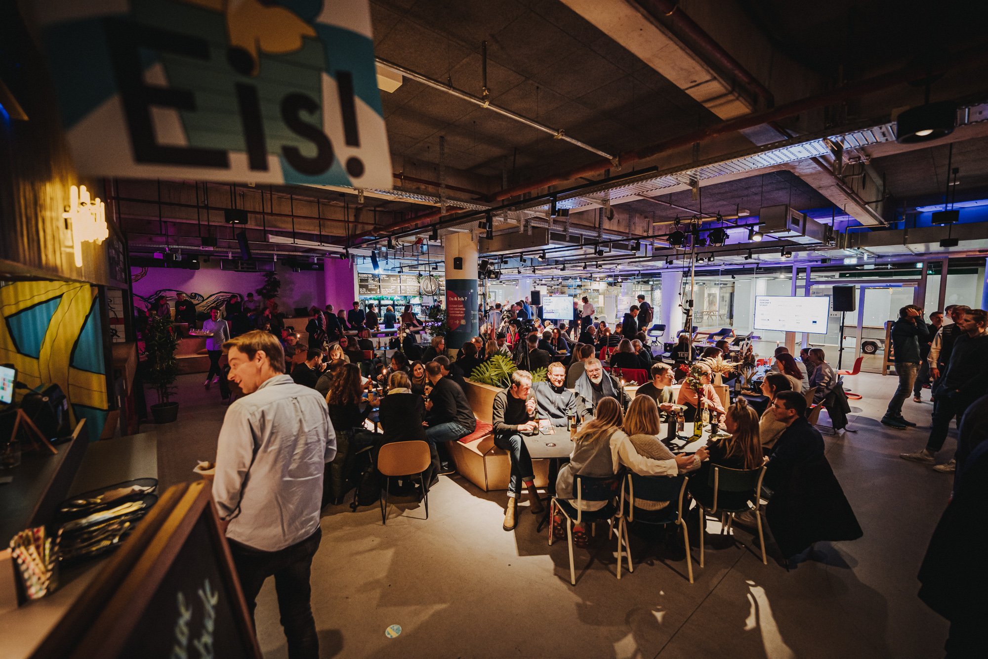 Menschen in einem modernen, gut beleuchteten Veranstaltungsraum, die an Tischen sitzen und sprechen, während auf Bildschirmen im Hintergrund Präsentationen angezeigt werden.