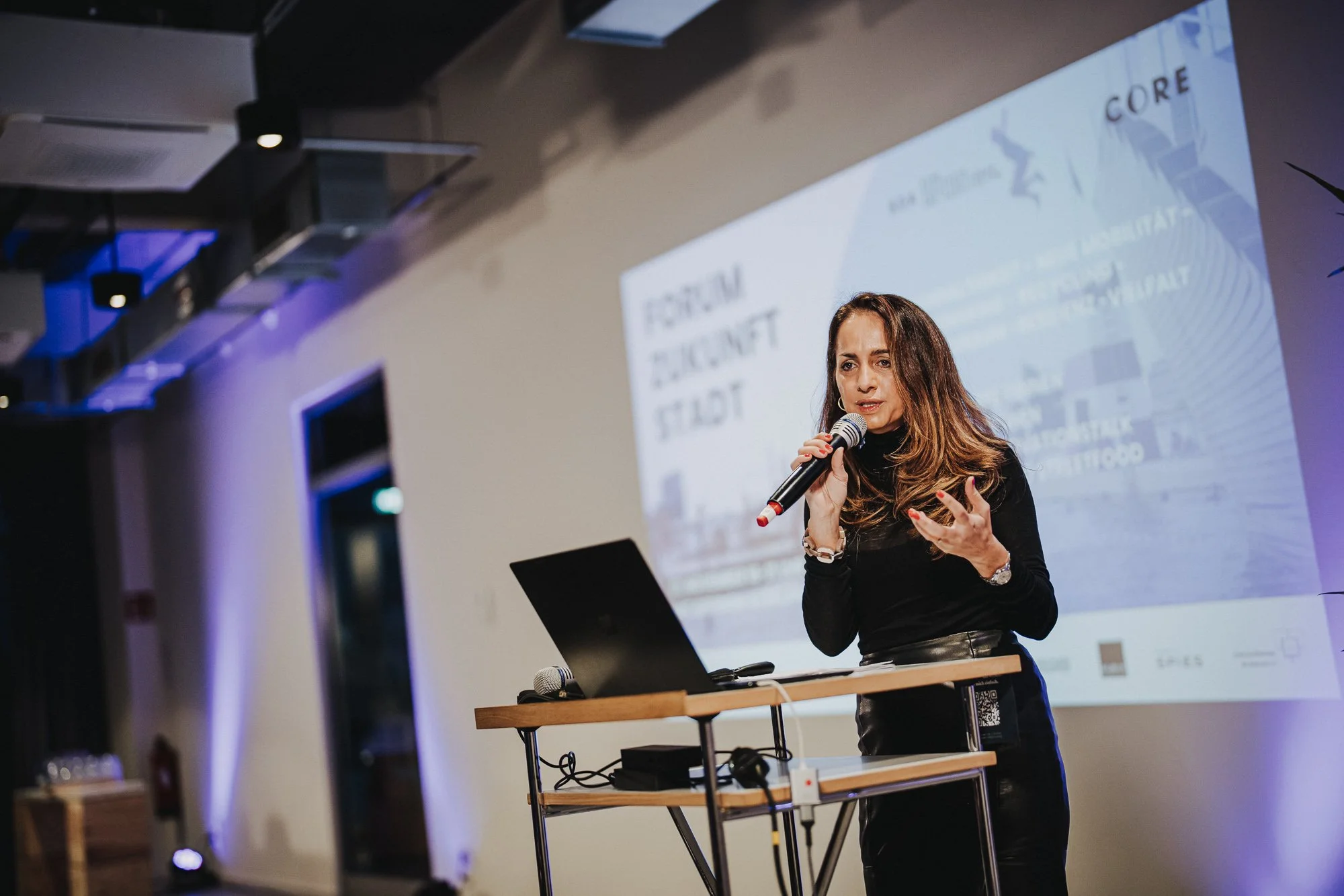 Eine Frau hält ein Mikrofon und spricht vor einem Projektor auf einer Bühne. Hintergrund zeigt eine Präsentation mit Text und Grafiken.