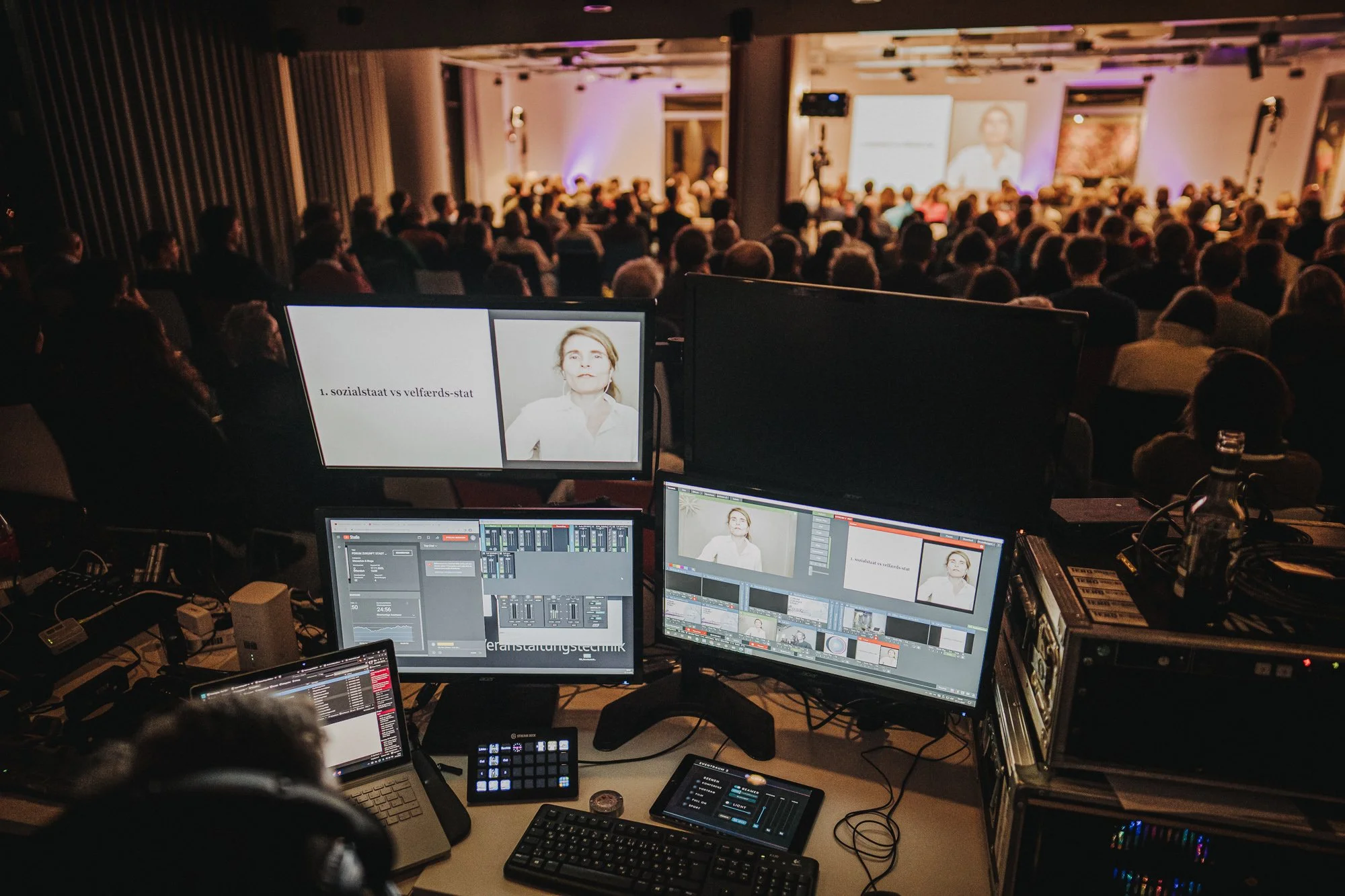 Blick auf eine Konferenz- oder Veranstaltungsszene mit einem Publikum im Hintergrund. Im Vordergrund arbeitet ein Technik-Team mit mehreren Monitoren, Laptops und Steuerungsequipment, um die Präsentation oder Videoaufnahmen zu steuern.