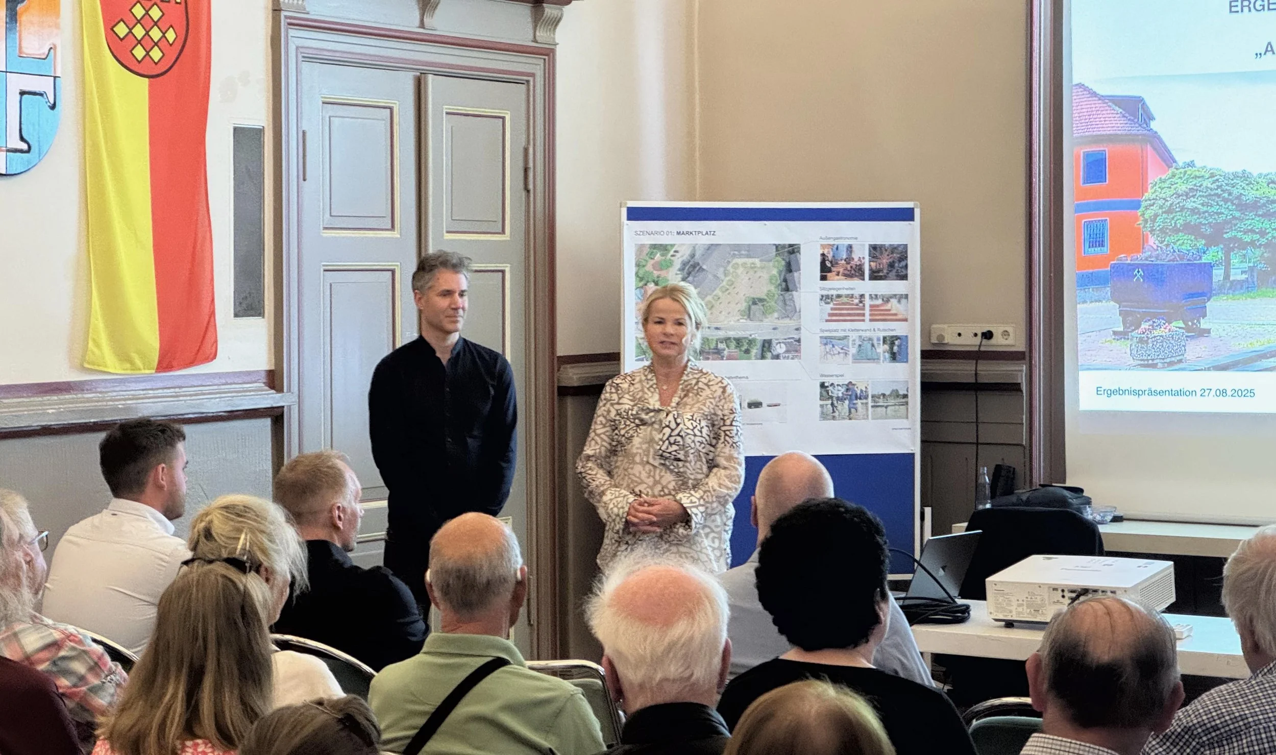 Workshop zum Projekt Aufwertung Marktplatz  Vienenburg. Bürgermeisterin von Vienenburg Frau Urte Schwerdtner und Lars Zimmermann von CITIES FOR FUTURE im Hintergrund. Beide stehen vor einem Publikum in einem Saal eines Gebäudes.