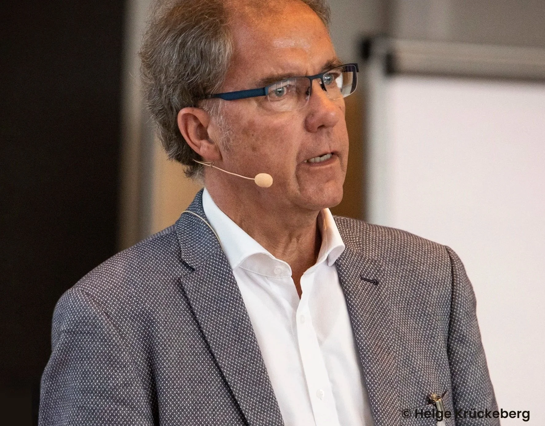 Klaus Mensing mit Brille und einem beige-braunen Anzug trägt ein Headset und spricht bei einer Präsentation. Klaus Mensing von CONVENT Mensing
