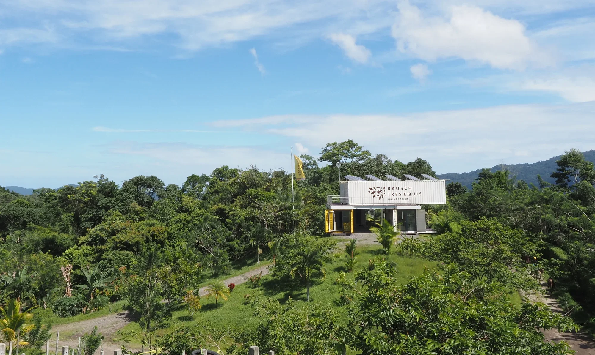 Ein modernes Gebäude auf einer grünen, bewaldeten Hügelkuppe, mit einem Schild, das 'Rausch Tres Equis' und 'Hacienda Cacao' liest, unter einem blauen Himmel mit Wolken.