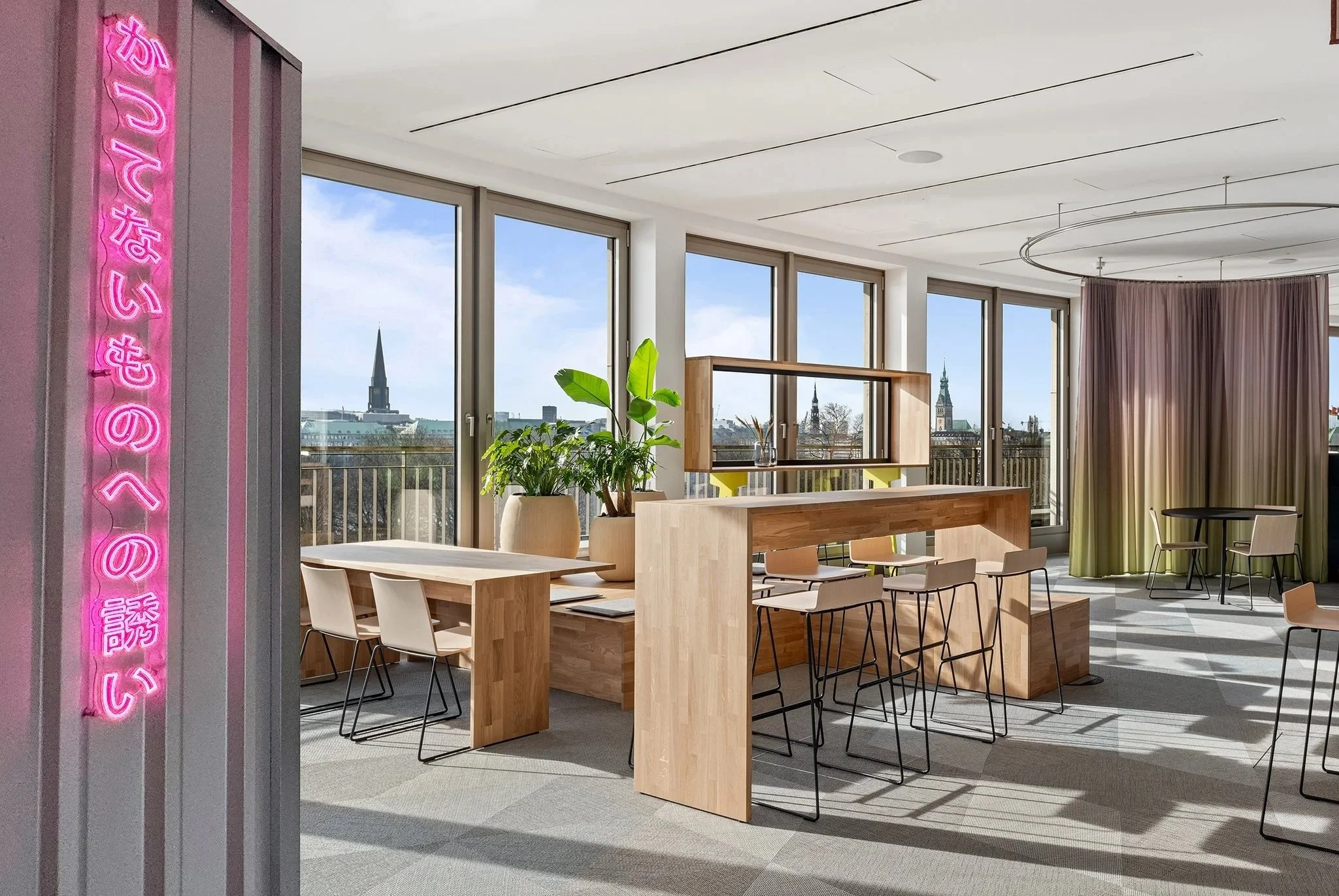 Dentsu Office: Raum mit Tischen und Stühlen, Pflanzen. Links ist der Blick durch Fenster auf Hamburg zu sehen. Themenbereiche: Interiorgestaltung, Innenarchitektur, Placemaking.