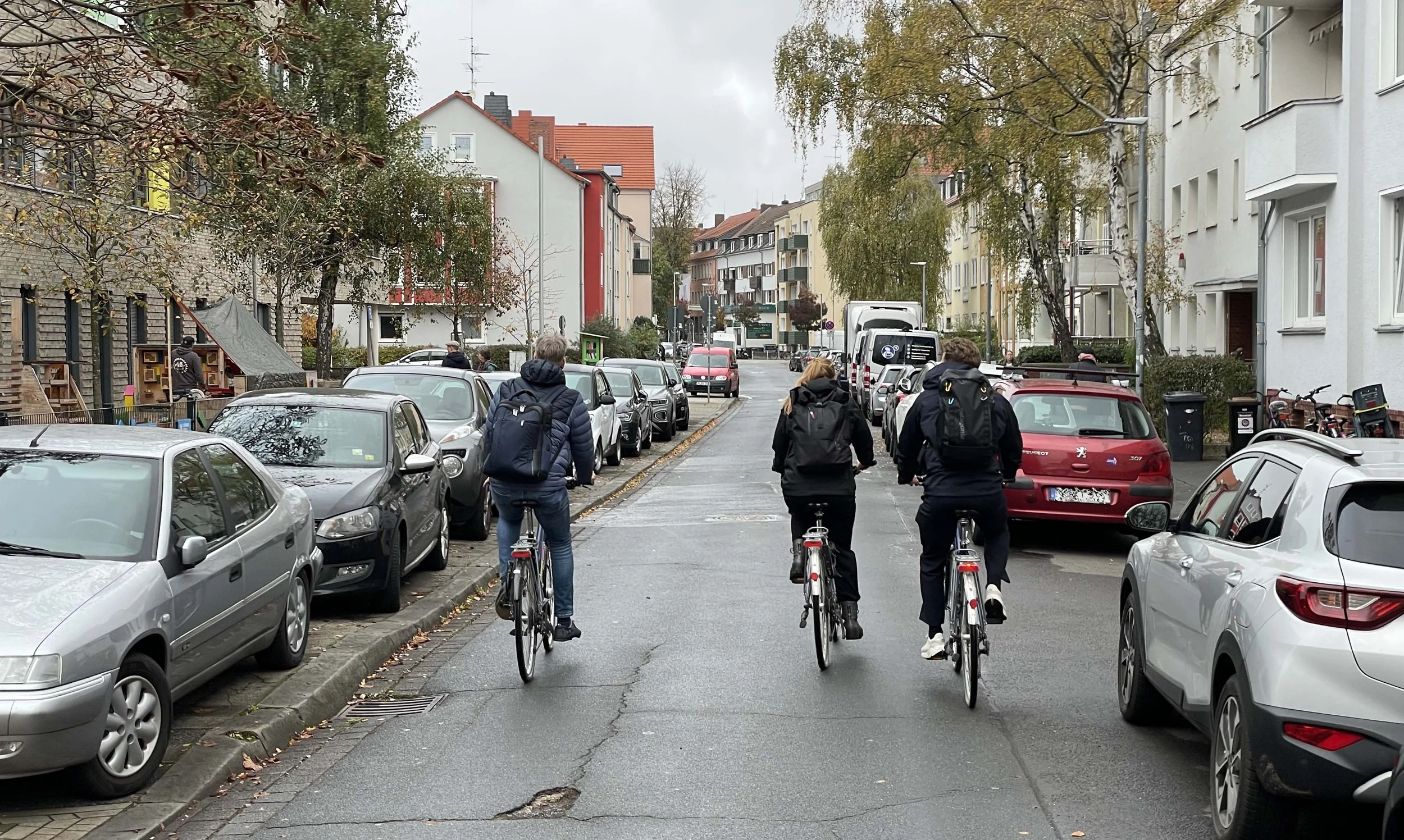 Foto einer Straße in Hannover mit drei Radfahrern von hinten. An den Seiten parkende Autos, Gebäude und Bäume.