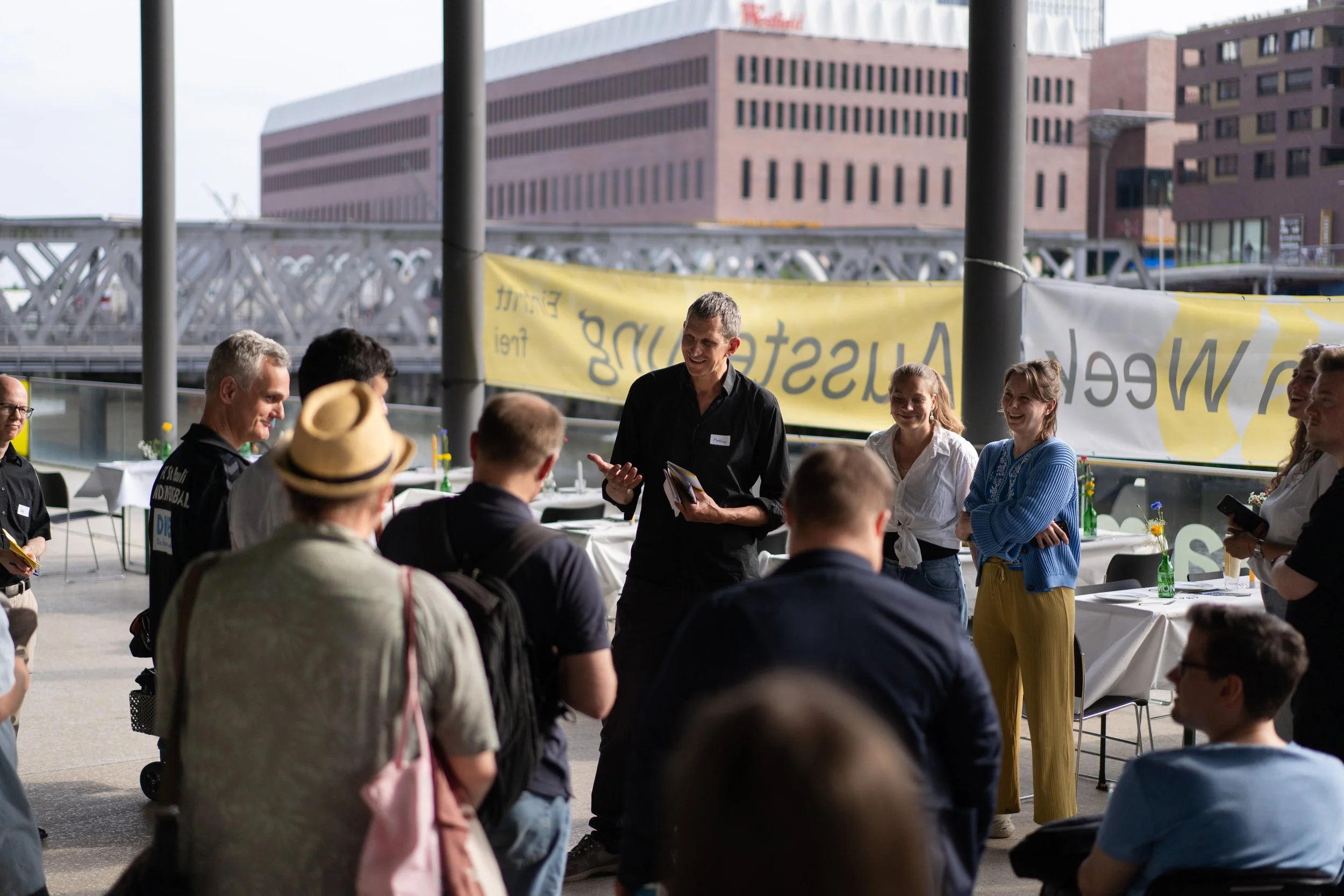 Gruppe von Menschen, die bei einer Veranstaltung im Freien zusammenstehen und einem Mann zuhören, der spricht, mit Tischen und Stühlen im Hintergrund und einer gelben Werbebanner.