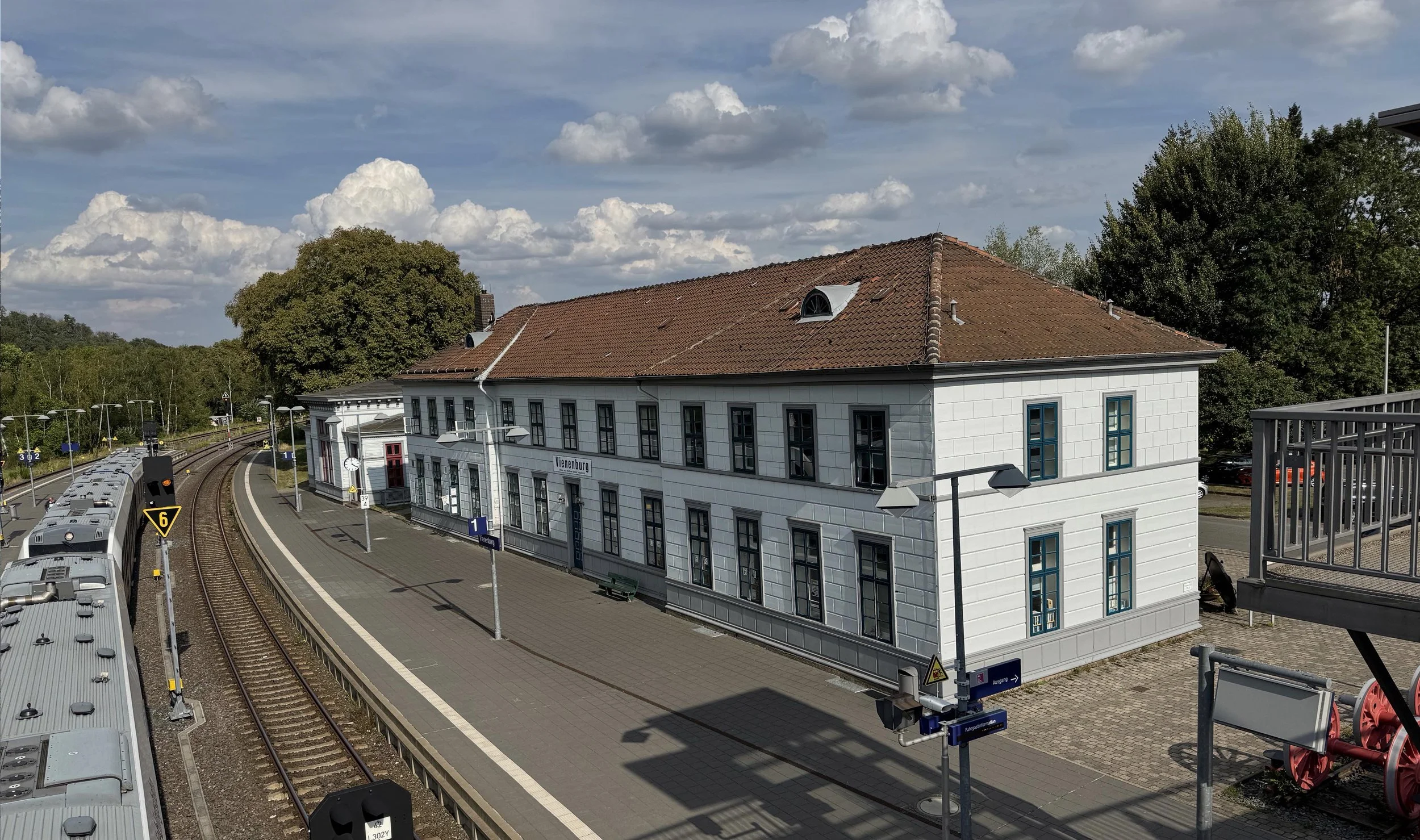 Foto Bahnhof Vienenburg. Aufgenommen von einer Brücke aus. Vorne links ein Zug von oben, dann ein leeres Gleis und ein Bahnsteig mit dem Bahnhof aus hellen Wänden und einem roten Dach.