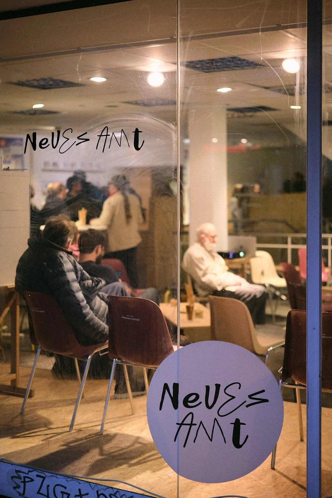 Menschen sitzen in einem Café oder Restaurant, im Hintergrund sind weitere Gäste und eine Bar sichtbar, auf der Scheiben steht 'Neues Amt Altona'.