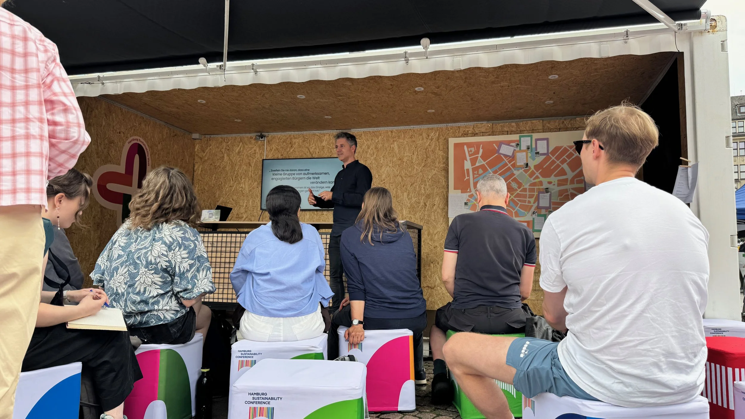 Lars Zimmermann von CITIES FOR FUTURE im Container als Bühne. Mehrere Menschen sitzen und stehen im Publikum.
Hamburg Sustainability Week 2026, Zukunftsgarten auf dem Rathausmarkt Hamburg.