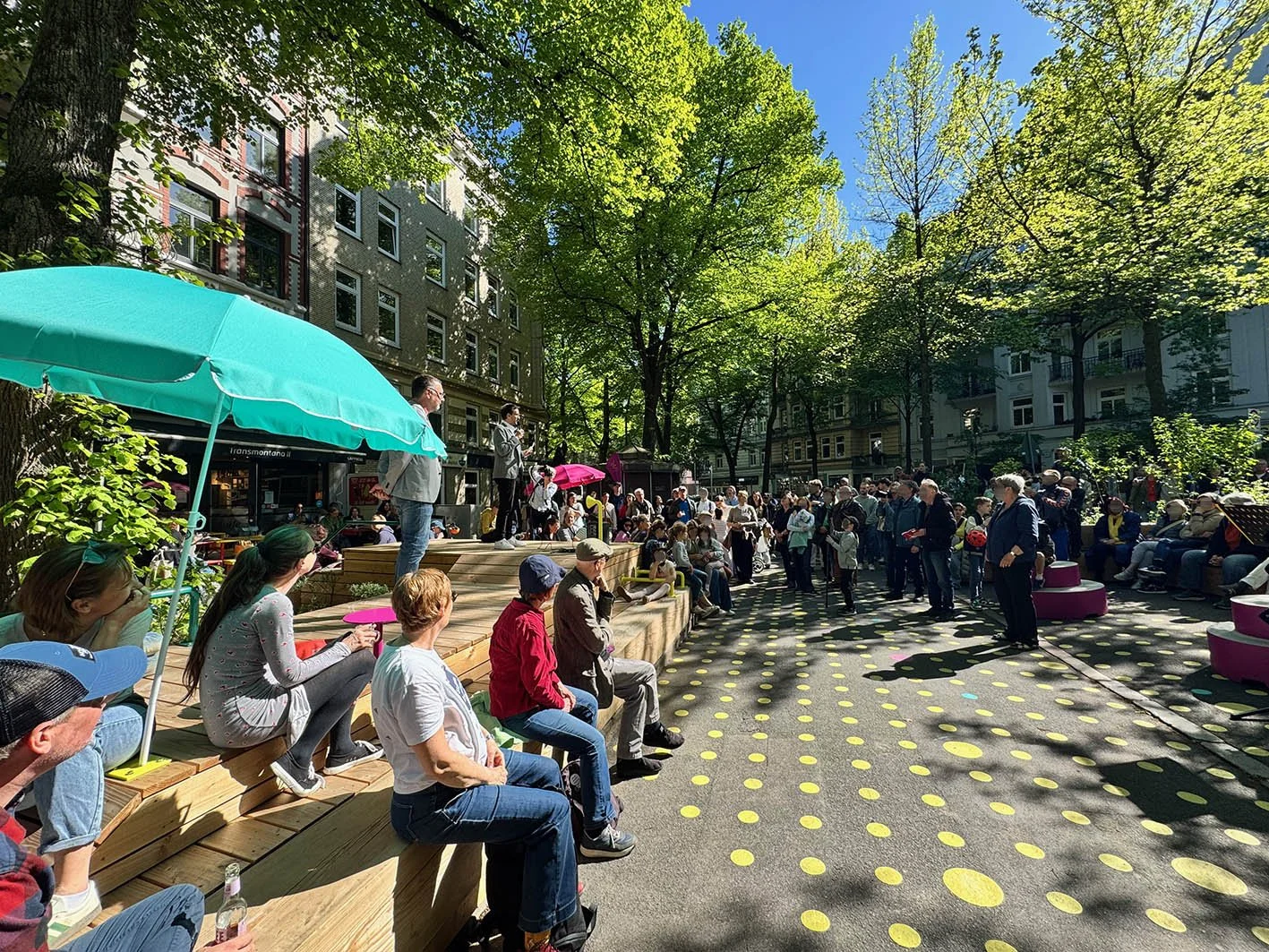 Eröffnung Parnass-Platz am 8. Mai 2026, viele Menschen hören einem Sprecher auf einer Holzbühne zu. Bunte Kreise auf der Straße, keine Autos.