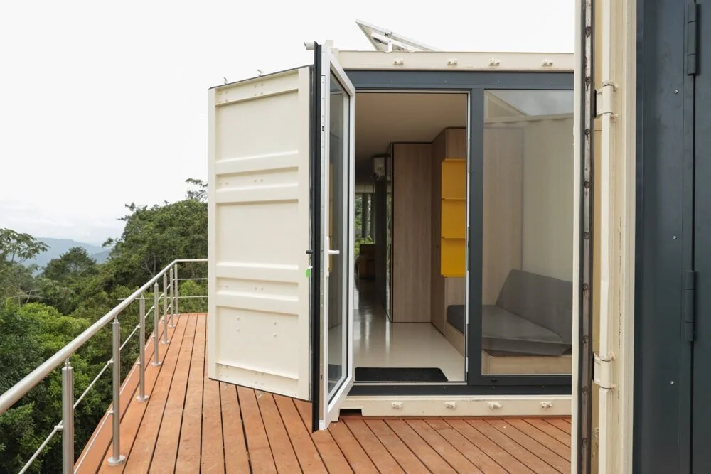 Eine Küsten- oder Waldkulisse mit einem kleinen Wohncontainer auf einem Holzdeck, mit offener Tür, Einblick in den Innenraum mit einer Sitzbank, Regal und Holzschränken.