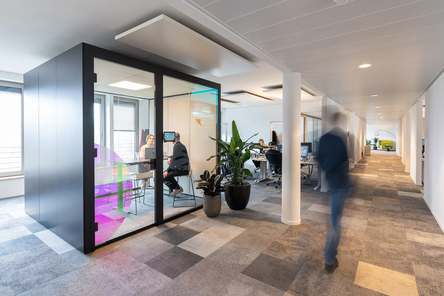Dentsu Office: Blick in einen Raum, der einen kleinen Raum bzw. eine Box beinhaltet. Dieser hat einen schwarzen Rahmen und an einer Seite bodentiefe Glasfenster und eine Glastür. Darin sitzen zwei Personen an einem Tisch und unterhalten sich.