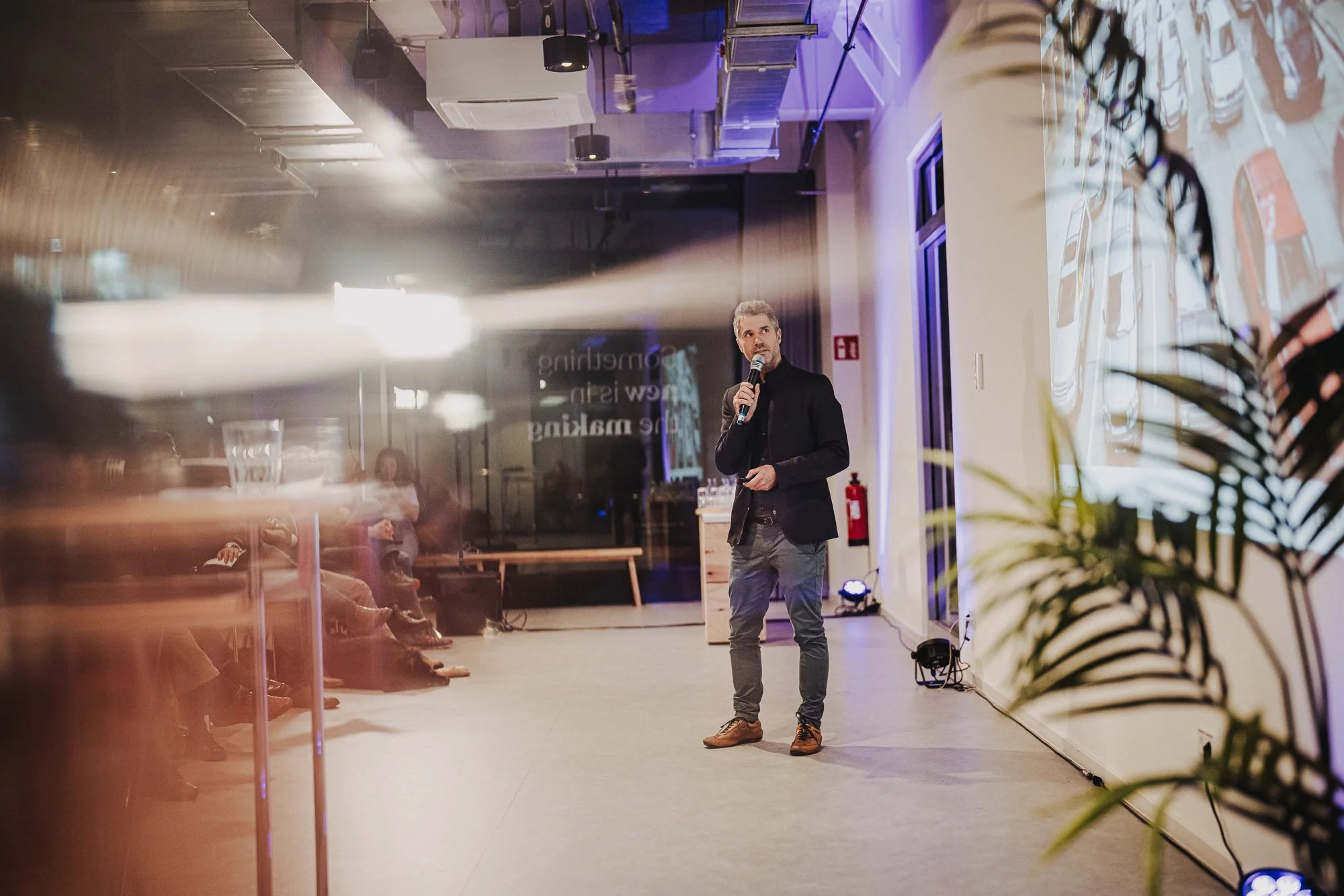 Ein Mann mit grauem Haar, schwarzer Jacke und grauen Hosen hält eine Hand am Körper und spricht auf einer Bühne in einem modernen Veranstaltungsraum mit Vorhang, Pflanzen und mehreren Zuschauern, die im Hintergrund sitzen. CORE Oldenburg